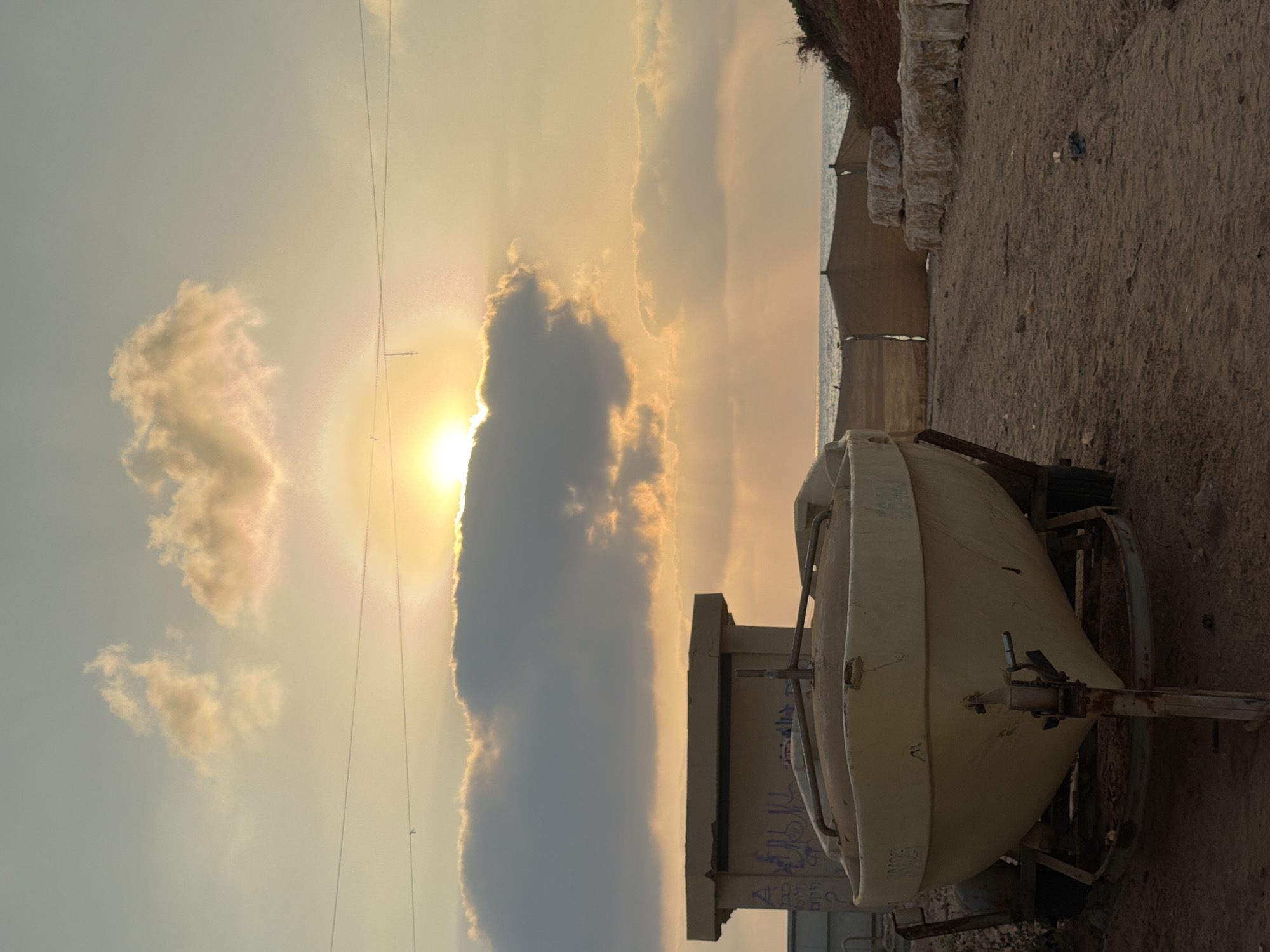 The image shows a boat on a trailer parked on a sandy area, likely near a beach. In the background, the ocean is visible under a cloudy sky with the sun setting or rising, casting a warm glow. There is a small structure with graffiti on it to the left of the boat. The location is identified as Hadera, Israel. The scene appears to be outdoors, with natural light illuminating the environment. There are no people visible in the image. The overall impression is one of a quiet, possibly abandoned or storage area near the coast.
