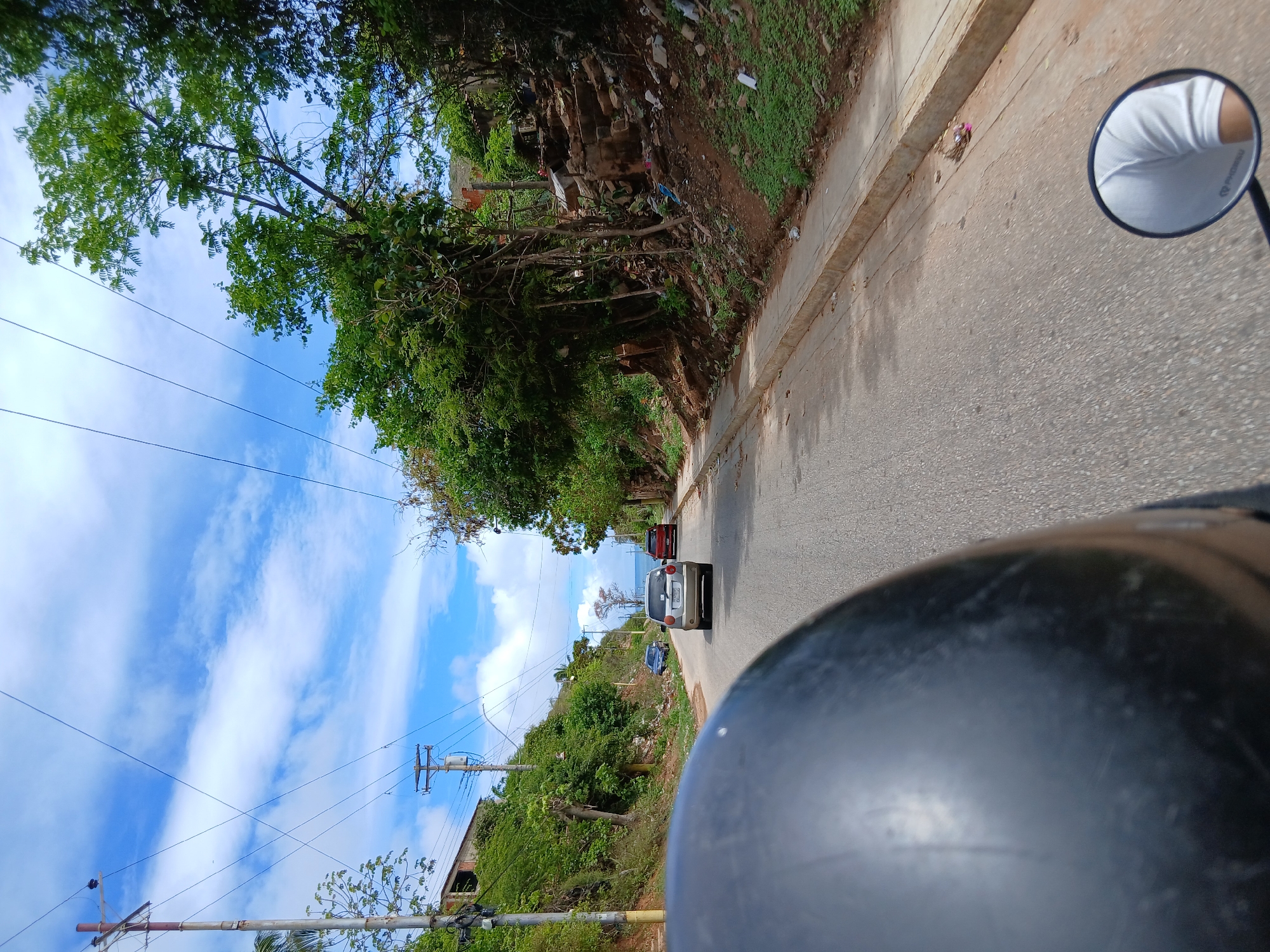 A first-person perspective shot from a motorcycle on a paved road, looking forward towards a slight uphill incline. In the foreground, the curved, dark surface of the motorcycle helmet dominates the lower left of the frame. To the right, a motorcycle's round rearview mirror reflects a portion of a person's white shirt and shoulder.
The road is bordered on the left by a steep, vegetated hillside with some scattered debris. A few houses or structures are visible on the upper part of this hill. On the right side of the road, a concrete sidewalk runs alongside a gentler, rocky incline covered with green vegetation and more debris.
Further down the road, several cars are visible. A silver hatchback is in the middle distance, followed by a red sedan. In the background, the road curves slightly to the right, and the sea and a distant horizon are visible under a bright blue sky with scattered white clouds. Utility poles and overhead wires are present along the road, indicating infrastructure. The overall impression is of a residential street in a coastal town, possibly in a developing area, with lush greenery and the proximity of the sea. The location is identified as Manzanillo, Venezuela.
