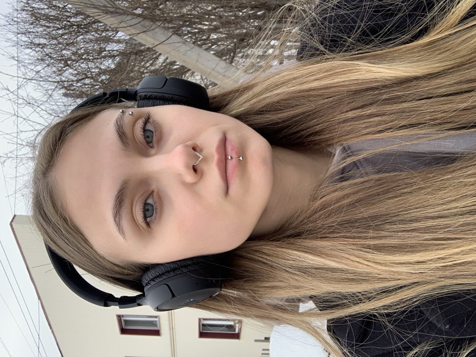 A close-up, waist-up portrait depicts a young adult female outdoors in the city of Фоки, Russia. The subject has light skin, light blue eyes, and long, straight, light brown hair parted in the middle. She maintains a neutral expression and looks directly at the camera. Her facial piercings include a silver-toned V-shaped septum ring with stones, a silver-toned barbell in her philtrum (medusa), a silver-toned vertical labret piercing with two spheres, and two silver-toned barbells on her left eyebrow (viewer's right). She wears black over-ear headphones, a light grey hoodie, and a dark outer jacket. The background features a cream-colored multi-story building with dark rectangular windows on the left and a cluster of bare-branched trees or bushes with a light-colored utility pole on the right. Patches of snow are visible on the ground in the lower left, indicating a cold weather environment under an overcast sky.
