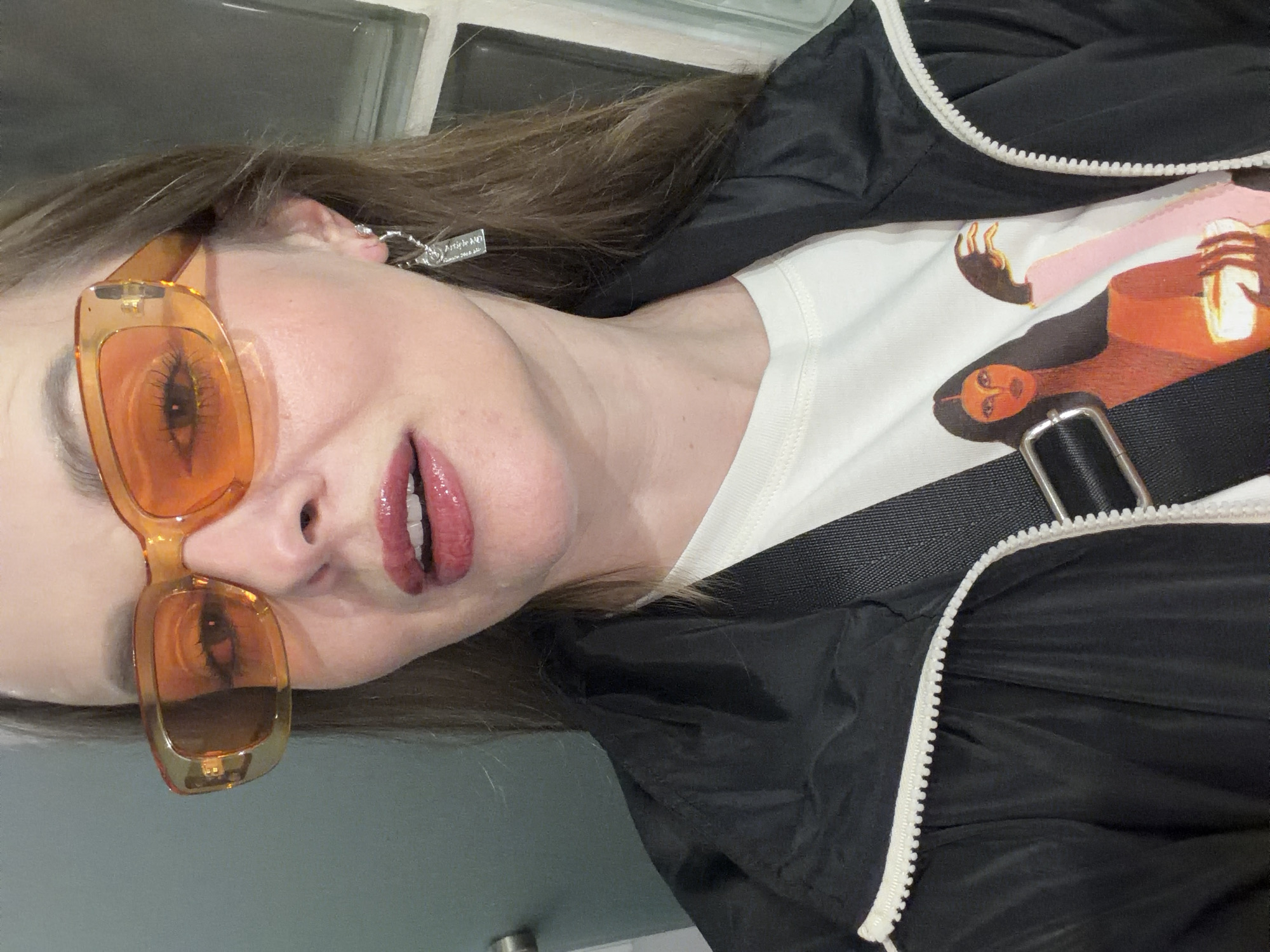 A woman is depicted at a tilted angle, wearing translucent orange glasses, glossy lipstick, a white t-shirt with a graphic image of a person, and a black jacket with white zippers. She also sports a silver earring. The location is not discernable from the image; the city of Moscow, Russia cannot be confirmed.