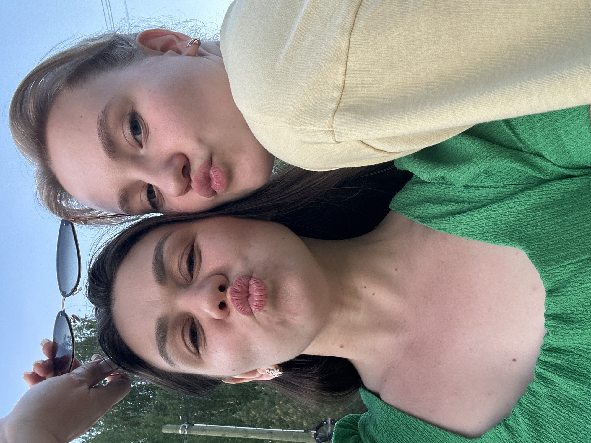 Two young women are posing close together for a selfie. Both are puckering their lips in a "kiss" expression. The woman in the bottom of the frame has dark brown hair, dark eyebrows, and is wearing a bright green textured shirt. She is holding a pair of sunglasses in her left hand. The woman at the top of the frame has blonde hair and is wearing a light yellow sweatshirt. The background is blurred and suggests an outdoor setting with some greenery and a glimpse of the sky. It is not possible to determine the exact location in Chaikovsky, Russia, from this image.