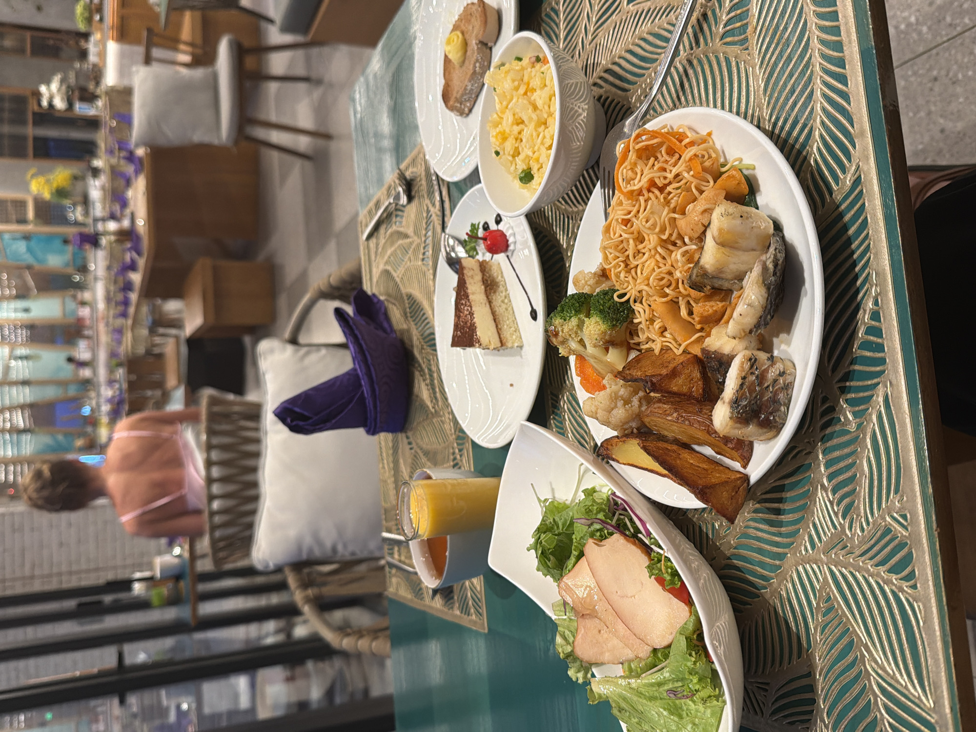This image shows a close-up view of a table laden with food and drinks in a restaurant or dining hall. In the foreground, there are several plates of food including a salad with lettuce and what appears to be sliced turkey or chicken, a plate with fried potato wedges, fish pieces, and noodles, a bowl of scrambled eggs, and a slice of tiramisu cake. A glass of orange juice and a cup of tea are also visible. A purple napkin is neatly folded on the table. In the background, a woman is seen from the back, wearing a pink top, sitting at a table. The restaurant appears to be spacious with tables and chairs arranged in the background, suggesting a buffet or dining area. The lighting suggests it might be evening. The overall scene depicts a meal setting in a well-lit indoor environment. The location is stated to be Dong Ninh Hoa Ward, Vietnam.