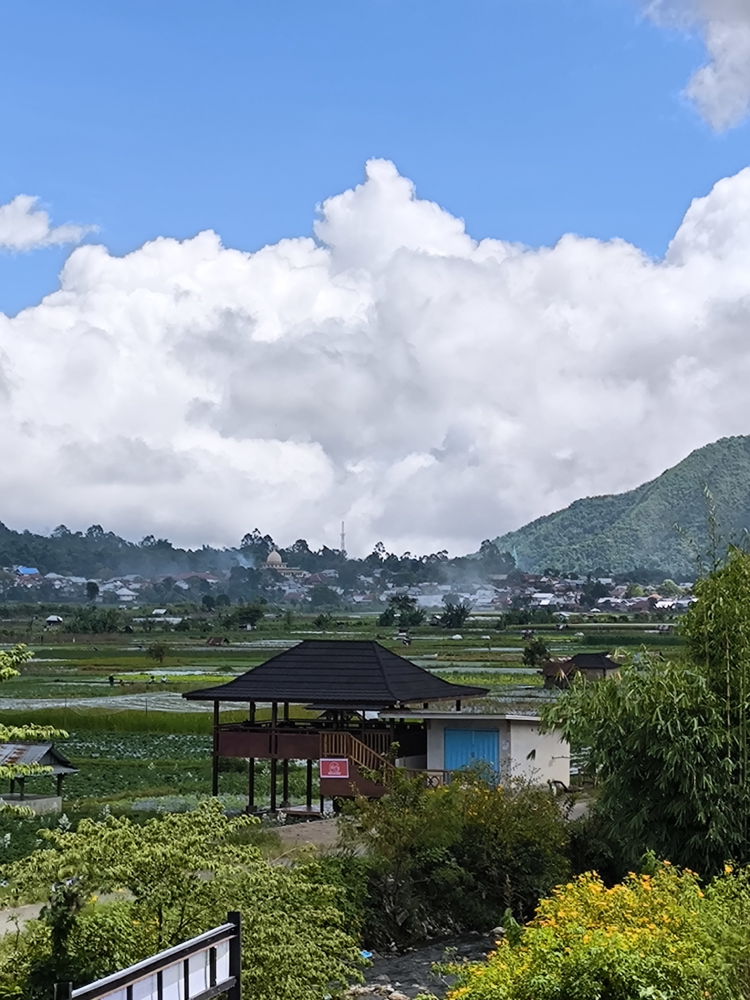 The image captures a scenic view in Sembalun Bumbung, Indonesia. In the foreground, a wooden pavilion with a black roof stands on stilts, adjacent to a small building with a blue door. Lush green foliage, including trees and flowering bushes with yellow blooms, fills the immediate foreground, partially obscuring a small stream.
The midground features rice paddies stretching towards a village nestled at the foot of a densely vegetated mountain. A mosque dome and minaret are visible within the village. The sky is filled with large, billowing white clouds against a clear blue backdrop. The overall scene depicts a tranquil landscape blending rural life with natural beauty.