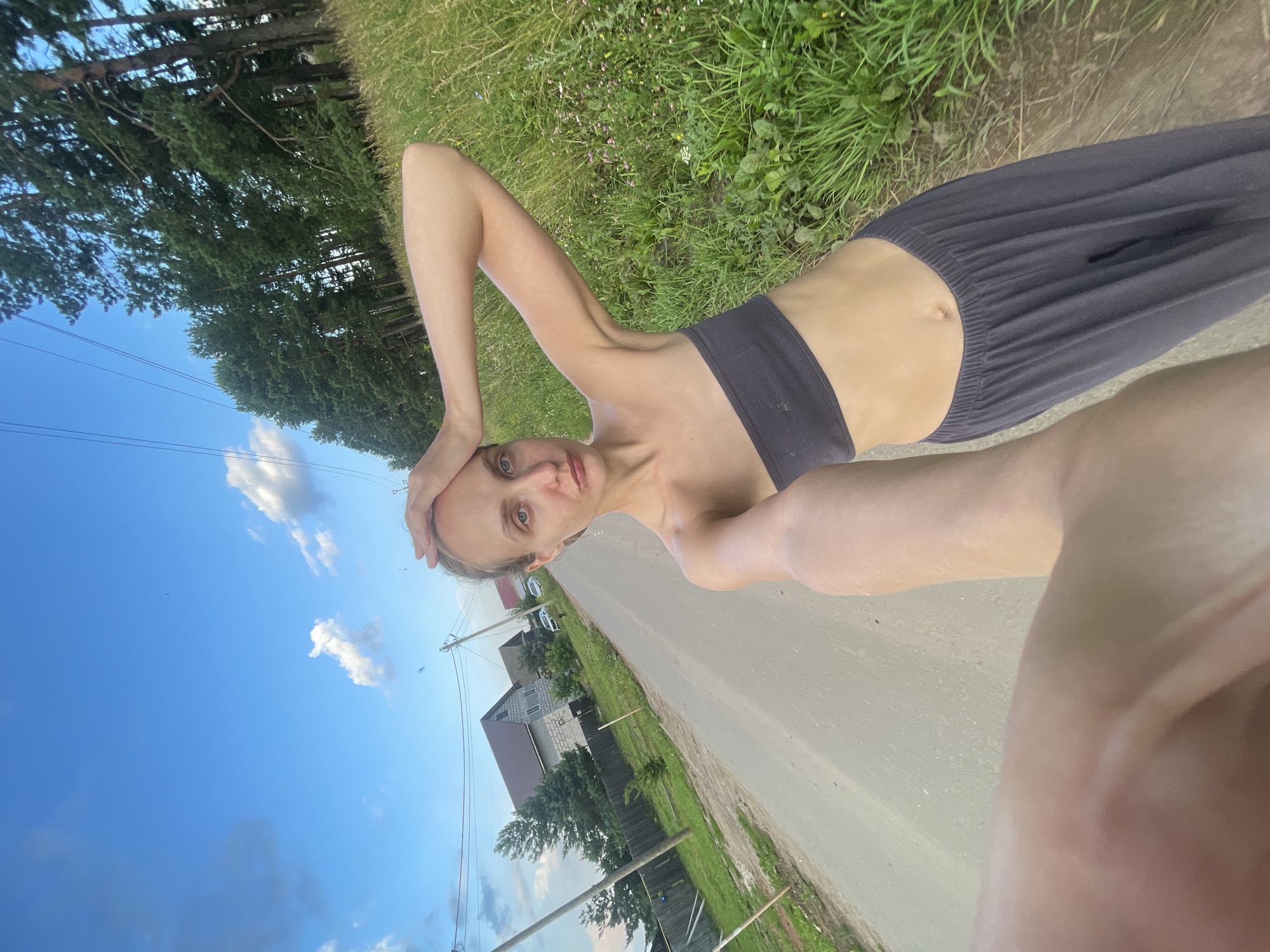 A young woman with light skin and blonde hair tied back, wearing a dark gray strapless top and dark gray loose-fitting pants, stands on a paved rural road. She holds her right hand to her forehead and her left arm is bent upward, with her hand near her head, as she takes a selfie. She has blue eyes and a neutral expression.
The road is unpaved and has some texture. To the left of the road, there are houses with dark roofs, a wooden fence, and some green trees. Overhead, power lines stretch across the scene, supported by wooden poles. To the right of the road, a field of tall green grass and wildflowers stretches towards a dense forest of pine trees. The sky above is a bright blue with scattered white clouds. The image appears to have been taken in the village of Podshivalovo, Russia, given the presence of rural housing and power lines typical of such areas. The lighting suggests it is late afternoon or early evening.