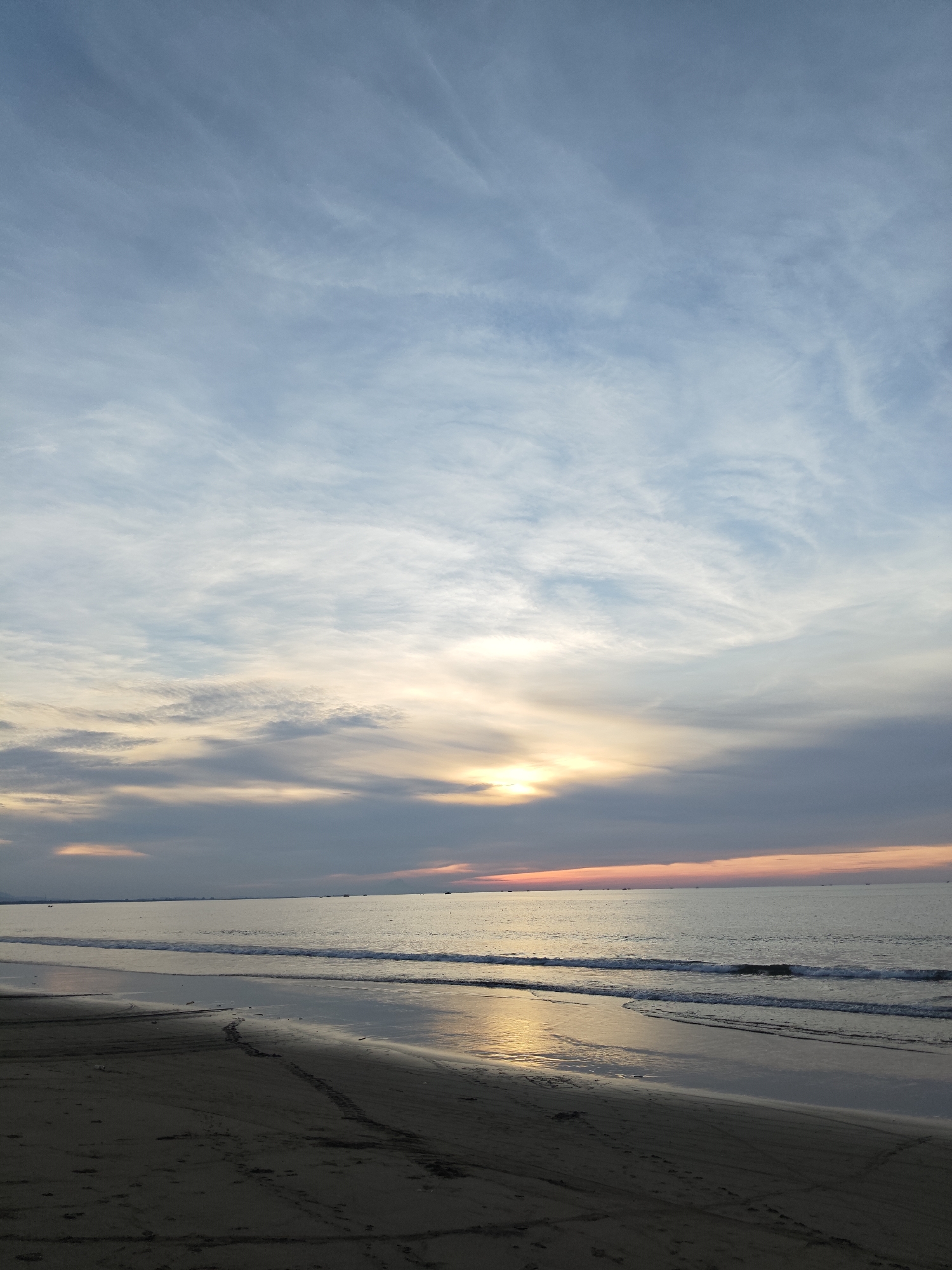 The image depicts a serene beach scene at what appears to be dusk or dawn. The sky is a canvas of soft blues, grays, and hints of orange and yellow near the horizon, suggesting the sun is either rising or setting. Wispy clouds stretch across the sky, reflecting the subtle colors of the sky. The ocean is calm, with gentle waves lapping at the shore. A thin line of boats is visible in the distance along the horizon. The foreground is a sandy beach, appearing dark and wet in places from the receding tide. Tire tracks or footprints are faintly visible on the sand. The overall atmosphere is peaceful and tranquil. The location is described as Curehtunong, Indonesia, indicating a coastal setting within that region.