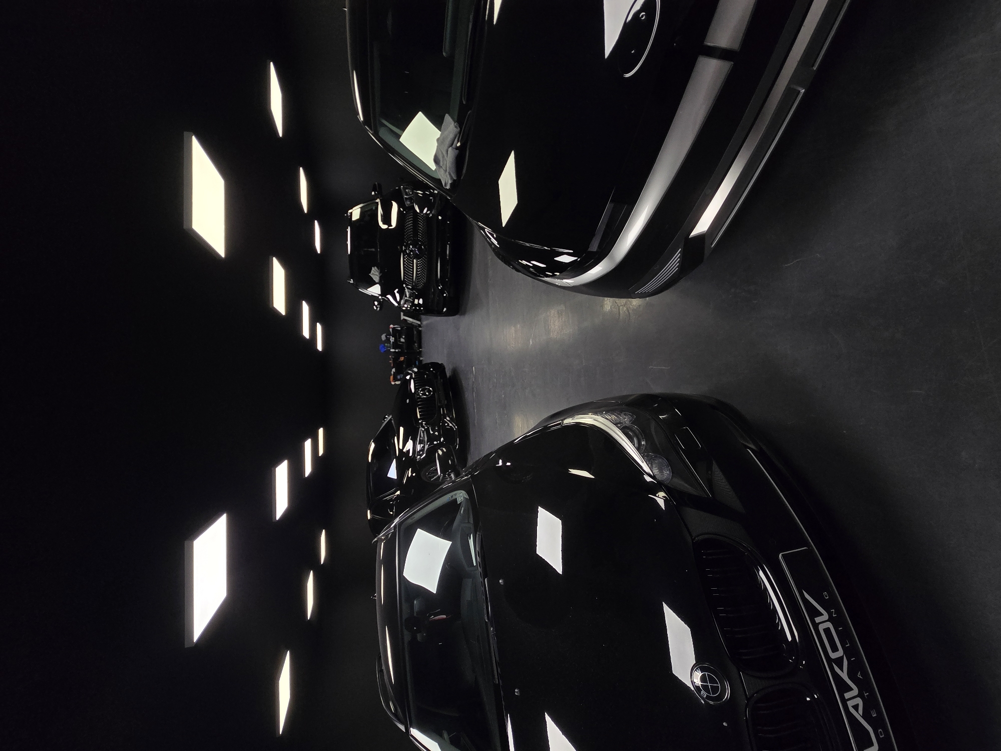 In a dimly lit garage, likely a car detailing studio, three black cars are parked. The car in the foreground left is a BMW sedan, its front end angled towards the viewer, revealing a polished grille and headlights. To its right, and slightly behind, is the front of a black car, possibly an electric vehicle, with a prominent silver stripe across its front. In the background, another black SUV, possibly a Mercedes-Benz, is visible. The ceiling is dark, with several recessed square and rectangular light panels illuminating the scene. The floor appears to be dark concrete or epoxy. The overall impression is one of a professional automotive detailing environment. The location is Ruse, Bulgaria.
