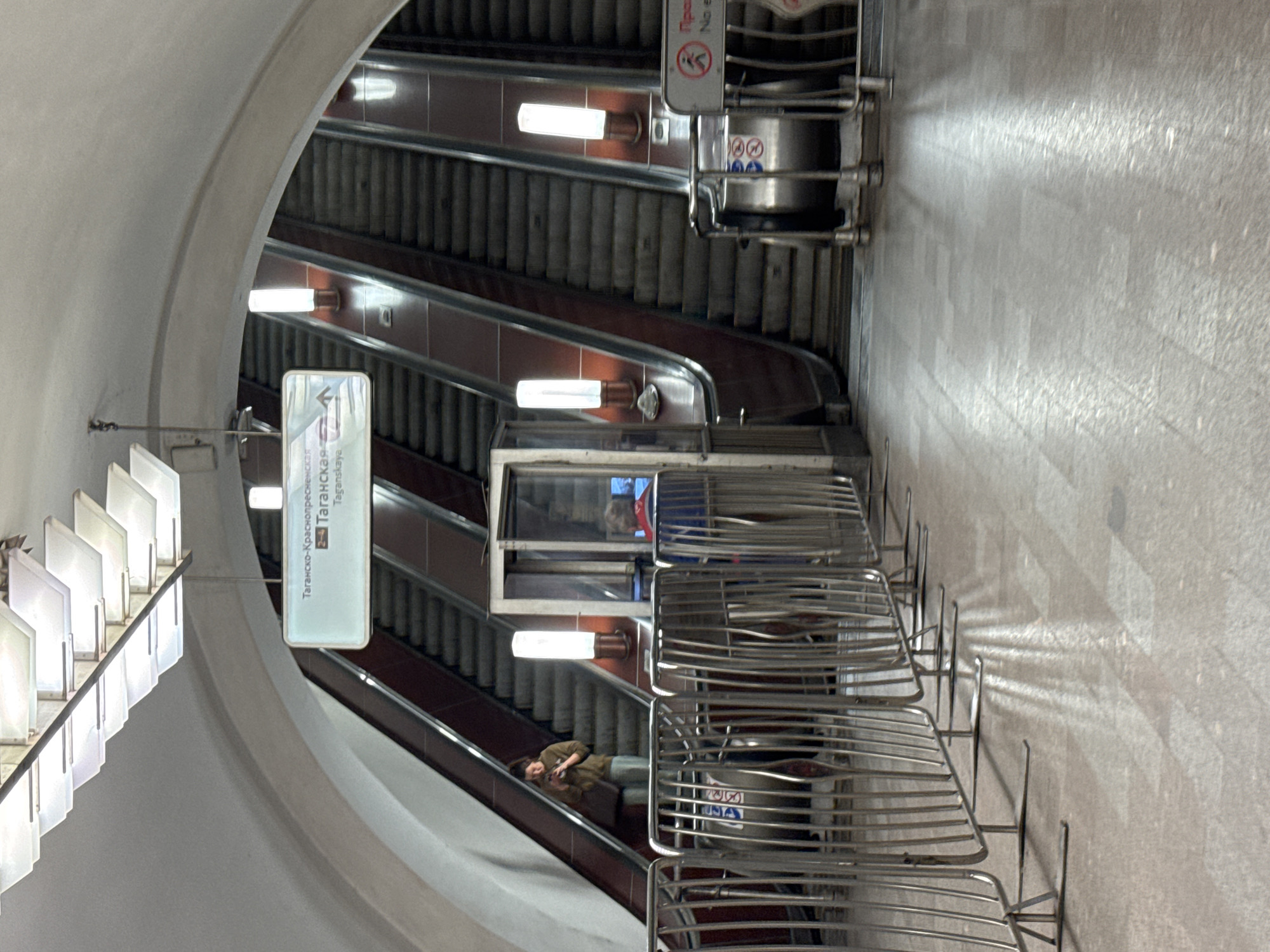 An eye-level, medium shot captures an underground metro station with multiple escalators. In the foreground, a row of metallic safety barriers extends across the frame, casting long shadows on the tiled floor. Behind the barriers, a woman stands on an escalator, looking at her phone. To her right, another escalator carries a person wearing a blue jacket and a red collar. In the background, a sign with Cyrillic text indicates "Taganskaya-Krasnopresnenskaya Line" and "Taganskaya." The station's architecture features a curved ceiling and modern lighting fixtures.