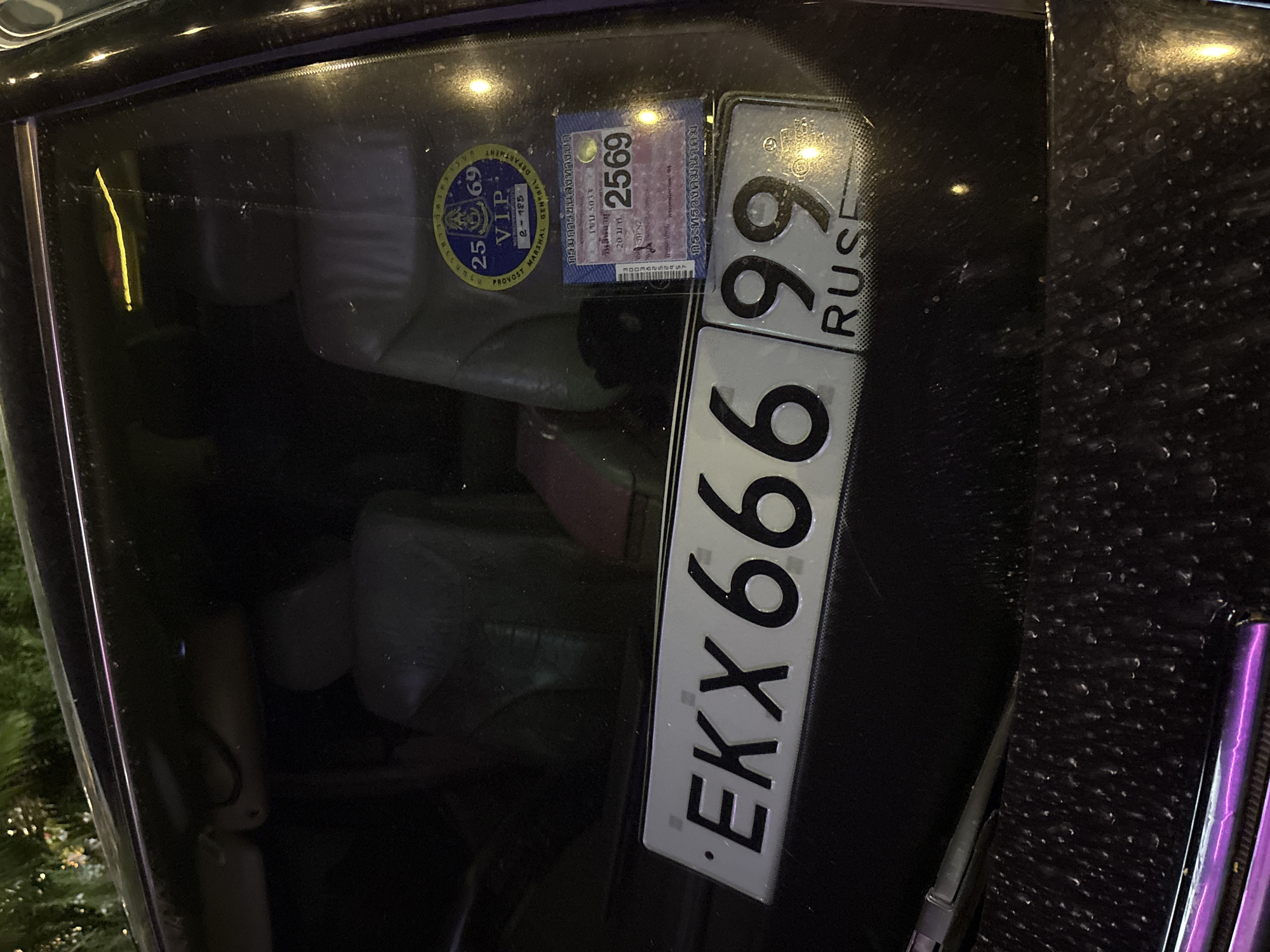 The image shows the interior of a car in Baan Bang Rak, Thailand. A circular sticker with "2569 VIP" and Thai writing is visible on the windshield. A rectangular sticker with the number "2569" and Thai writing is also present. Next to these stickers is a vertical license plate reading "EKX 666 99 RUSE". The car seats are visible in the background. The exterior of the car shows a dark, textured surface, likely carbon fiber.