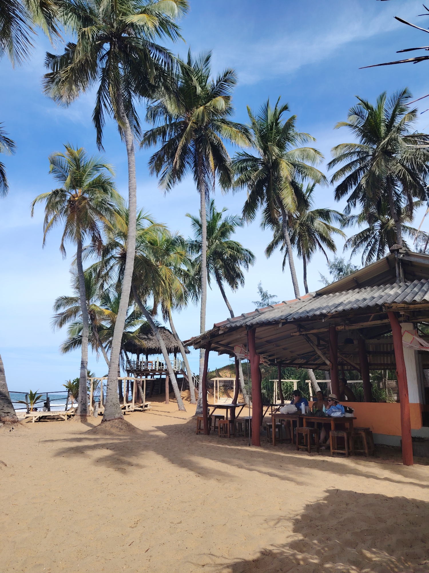 This image depicts a beach scene with several palm trees under a bright blue sky with scattered clouds. In the foreground, there is a sandy beach with shadows cast by the palm trees. To the right, a rustic open-air structure with a tiled roof serves as a seating area. Several wooden tables and stools are visible, and at one table, two people are seated. In the background, a raised wooden structure, possibly a deck or platform, can be seen among the trees, and a section of the ocean is visible in the distance. The overall impression is a relaxed, tropical beach resort or cafe.