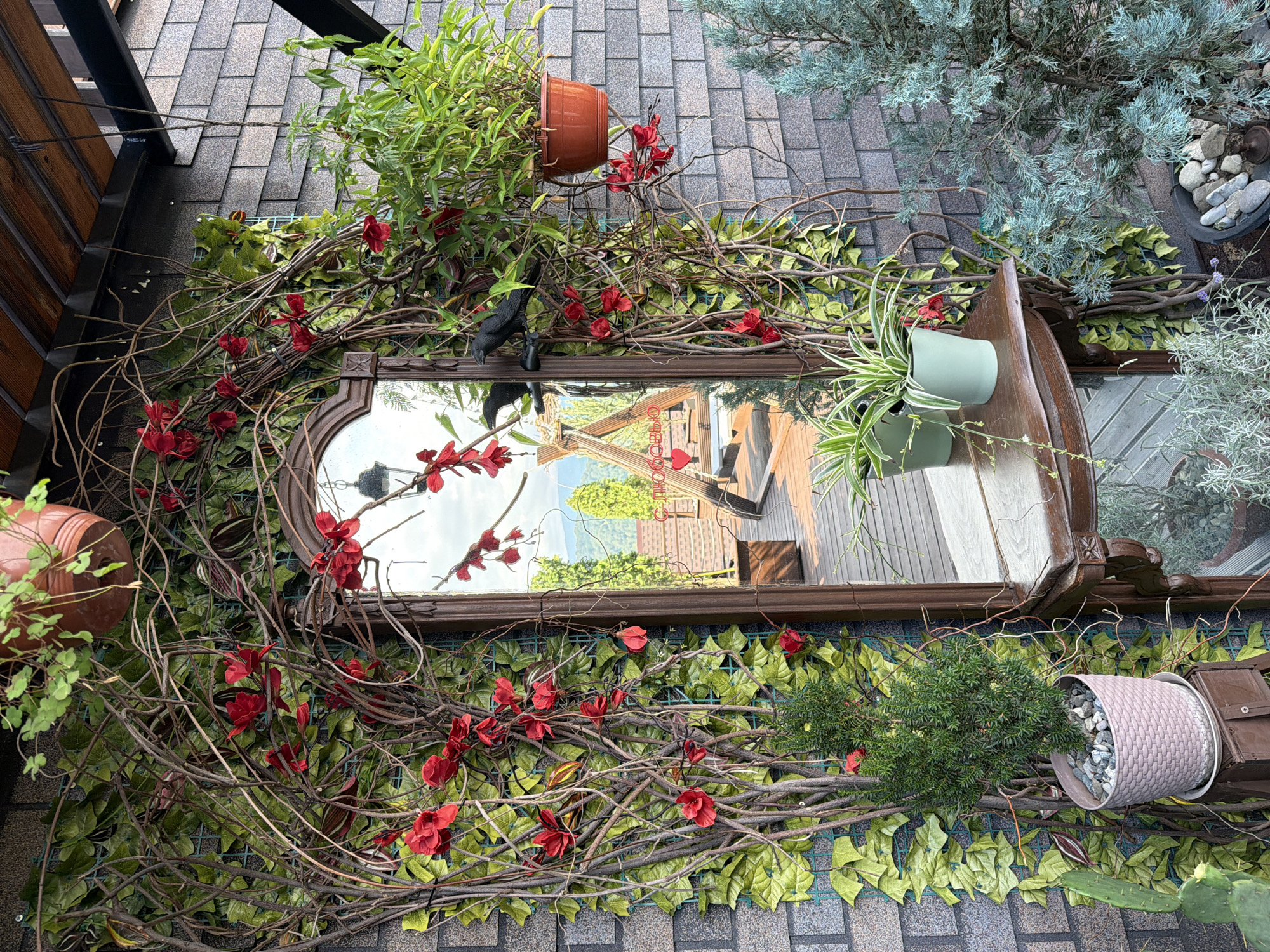 In Nizhnyaya Shilovka, Russia, a decorative outdoor scene features a wooden-framed mirror surrounded by artificial vines and red artificial flowers, attached to a dark grey brick wall. A black crow figurine is perched on the left side of the mirror's vine decoration. Two potted plants, one with green leaves in a light blue pot and another with striped leaves in a sage green pot, sit on a wooden shelf below the mirror. To the right of the mirror, a larger potted plant with long green leaves hangs from a terracotta pot. In the foreground, a small evergreen plant in a pink textured pot is positioned on a dark wooden stand. A cactus plant is visible to the left, and a blue-green conifer tree stands to the right, with a small pot filled with stones in front of it. The mirror reflects a wooden deck, a swing set, and a mountainous landscape under a cloudy sky. The scene is covered by a wooden roof structure with visible beams.
