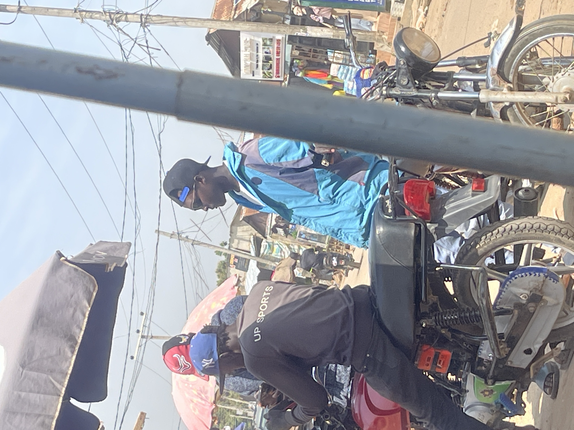 This is a photograph taken in an outdoor market environment in Abuja, Nigeria. The image is partially obscured by a thick, gray pole in the foreground, running vertically down the right side of the frame. In the midground, two young men are seated on a red motorcycle. The man on the left wears a dark grey sweatshirt with "UP SPORTS" written in white lettering across the back, a blue New York Yankees baseball cap, and dark pants. He is holding a smartphone in his left hand. The man on the right is wearing a multi-colored blue and purple jacket over a white t-shirt, a black baseball cap, and sunglasses. He appears to be smoking. Both men are facing to the left of the frame.
Behind the motorcycle, the scene opens up to a bustling market. There are several stalls and buildings visible, with corrugated metal roofs and wooden structures. A pink and red umbrella is visible behind the men on the motorcycle. In the background, another motorcycle carrying a rider is seen moving away from the camera. Power lines and utility poles crisscross the sky. The ground appears to be a dirt or unpaved road. The overall atmosphere suggests a vibrant, everyday scene within a Nigerian urban market.