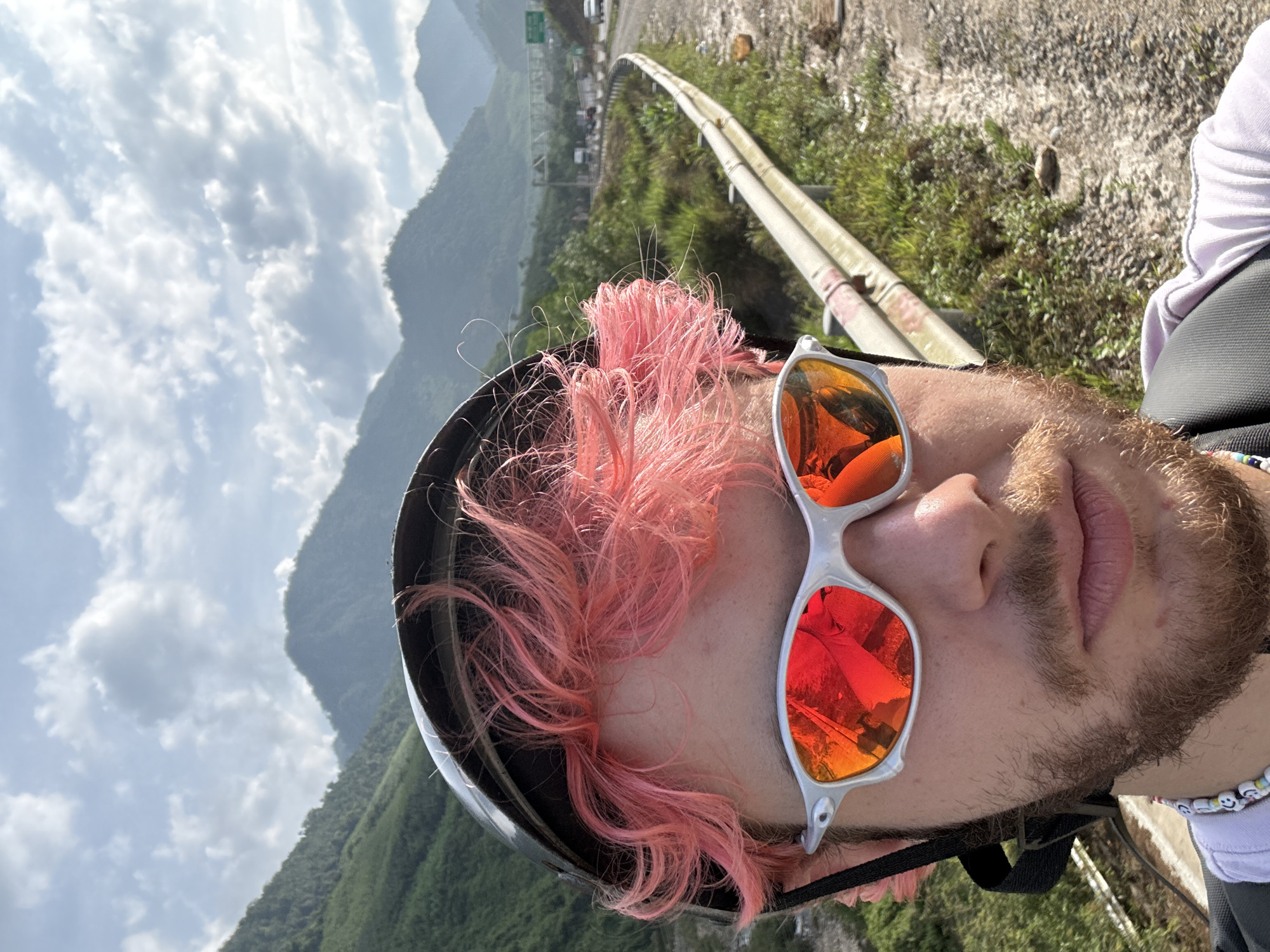 A selfie taken by a person with pink hair, wearing a helmet and sunglasses, is oriented vertically, showcasing a mountainous landscape in the background. The sunglasses reflect a red-orange image, possibly of the person taking the photo or another element in the scene. The person is likely positioned near a road with a metal guardrail. The background reveals a lush, green mountainous region under a sky with scattered clouds.
While the exact location in Đà Nẵng is difficult to determine from the image alone, the mountainous backdrop suggests the person is likely near or traveling along one of the roads that leads through the mountains outside the city, possibly towards locations like Ba Na Hills or the Hai Van Pass.