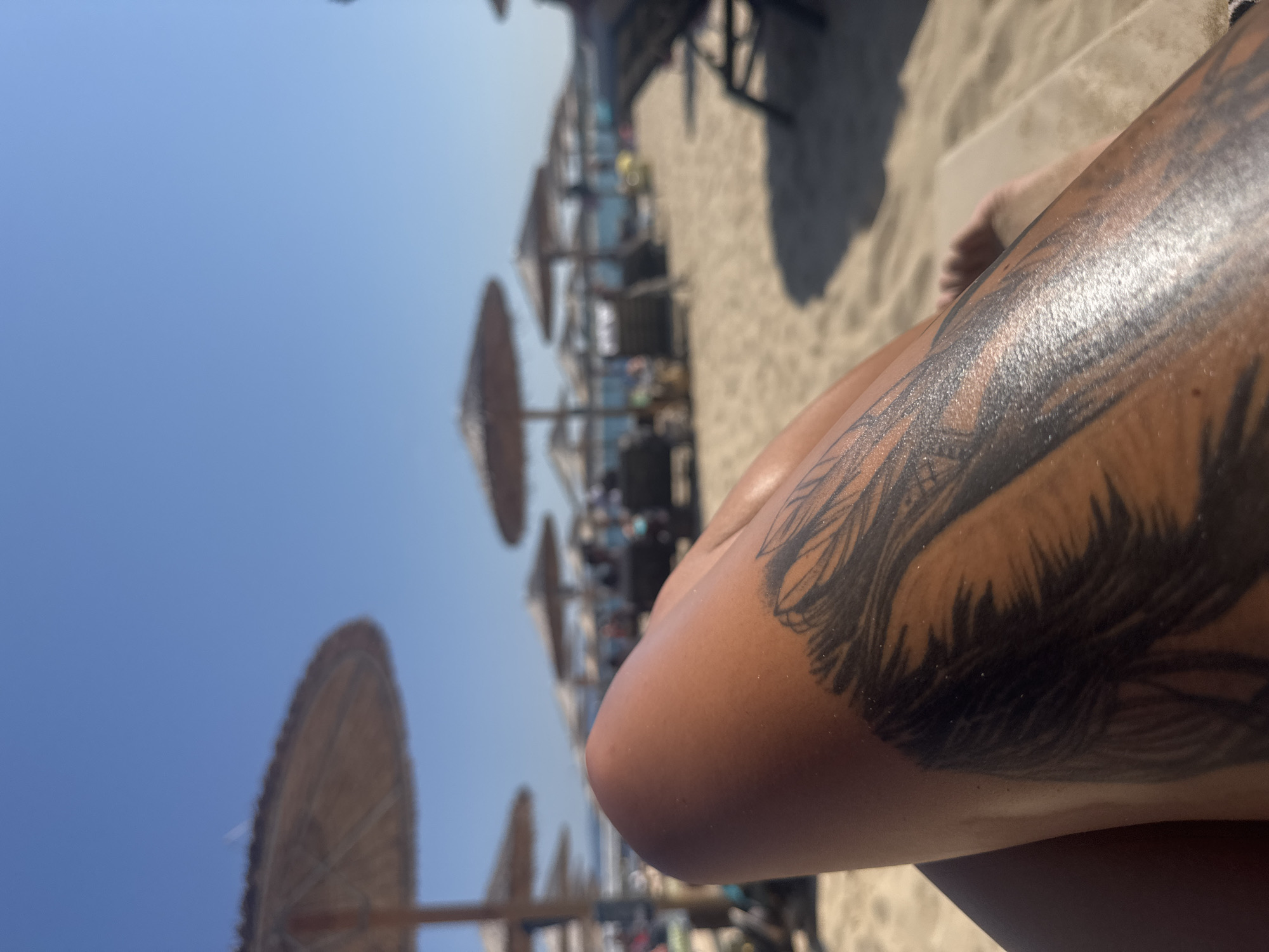 The image depicts a close-up of a person's tanned leg with a tattoo of what appears to be feathers. The leg is resting on a beach chair, suggesting a leisure activity. In the background, a beach scene unfolds under a clear blue sky. Numerous thatched umbrellas provide shade to empty beach chairs, indicating a relaxed atmosphere. The sandy beach extends towards the distant sea, which is a calm blue. The overall impression is one of a serene summer day at a coastal resort. The location is identified as Primorsko, Bulgaria, a well-known Black Sea resort town famous for its beaches and natural beauty.