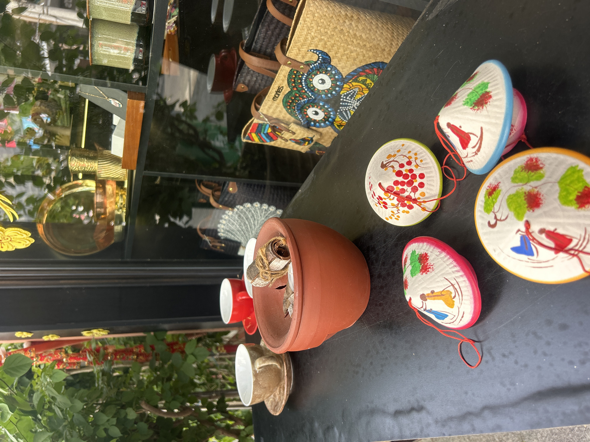 A black table next to a glass display in Nha Trang, Vietnam, displays various items. Five painted Vietnamese conical hats with red strings lie on the table. A brown terracotta pot with dried plant material sits next to the hats, along with several teacups. The glass display behind the table contains jewelry, purses, and other decorative items. Plants are visible outside the glass display.