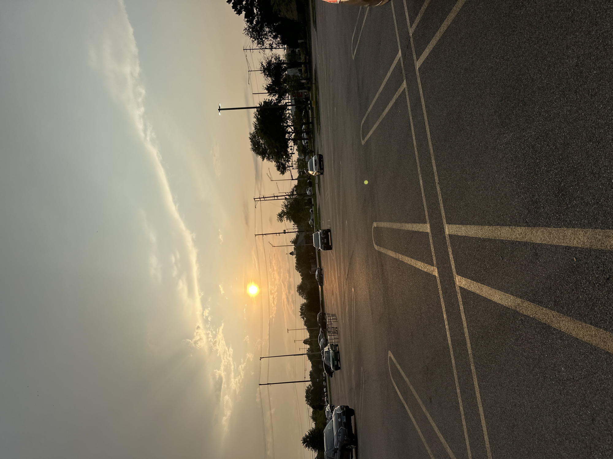 The image depicts a view of a nearly empty outdoor parking lot at dusk. The sun, a bright orange orb, is visible in the sky, casting a warm glow and creating visible sunbeams. The sky is a mix of hazy clouds and clear patches. In the distance, trees and utility poles are silhouetted against the sky. Several cars are parked and a few are in motion, driving through the lot. The foreground is dominated by the asphalt of the parking lot, marked with yellow lines indicating parking spaces. The scene suggests a commercial or retail area, possibly a shopping center, located in Schaumburg, United States of America, based on the context of similar images. The overall impression is of a quiet evening in a suburban environment.