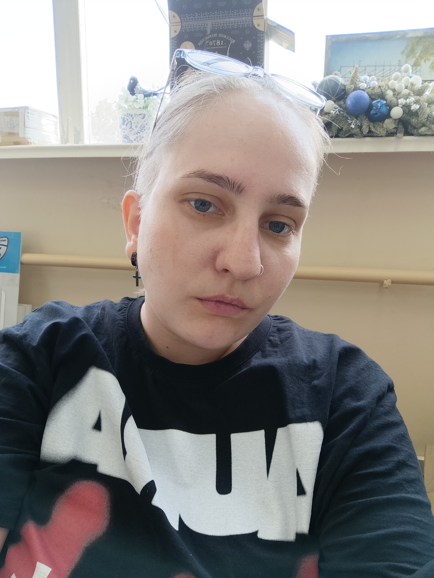 A close-up, eye-level shot of a young person with light skin and short, light blonde hair, their blue eyes looking directly at the camera. They have a nose ring and wear a black t-shirt with large, white lettering. A small cross earring is visible in their left ear.
Behind the person, a shelf is visible with various objects. In the background, daylight streams in through a window, illuminating a floral arrangement with blue and white ornaments. A stacked box and a framed painting sit on the shelf behind the floral arrangement. The top of a blue and gold box can be seen to the left of the painting.
There is no indication that this image was taken in Chaikovsky, Russia.