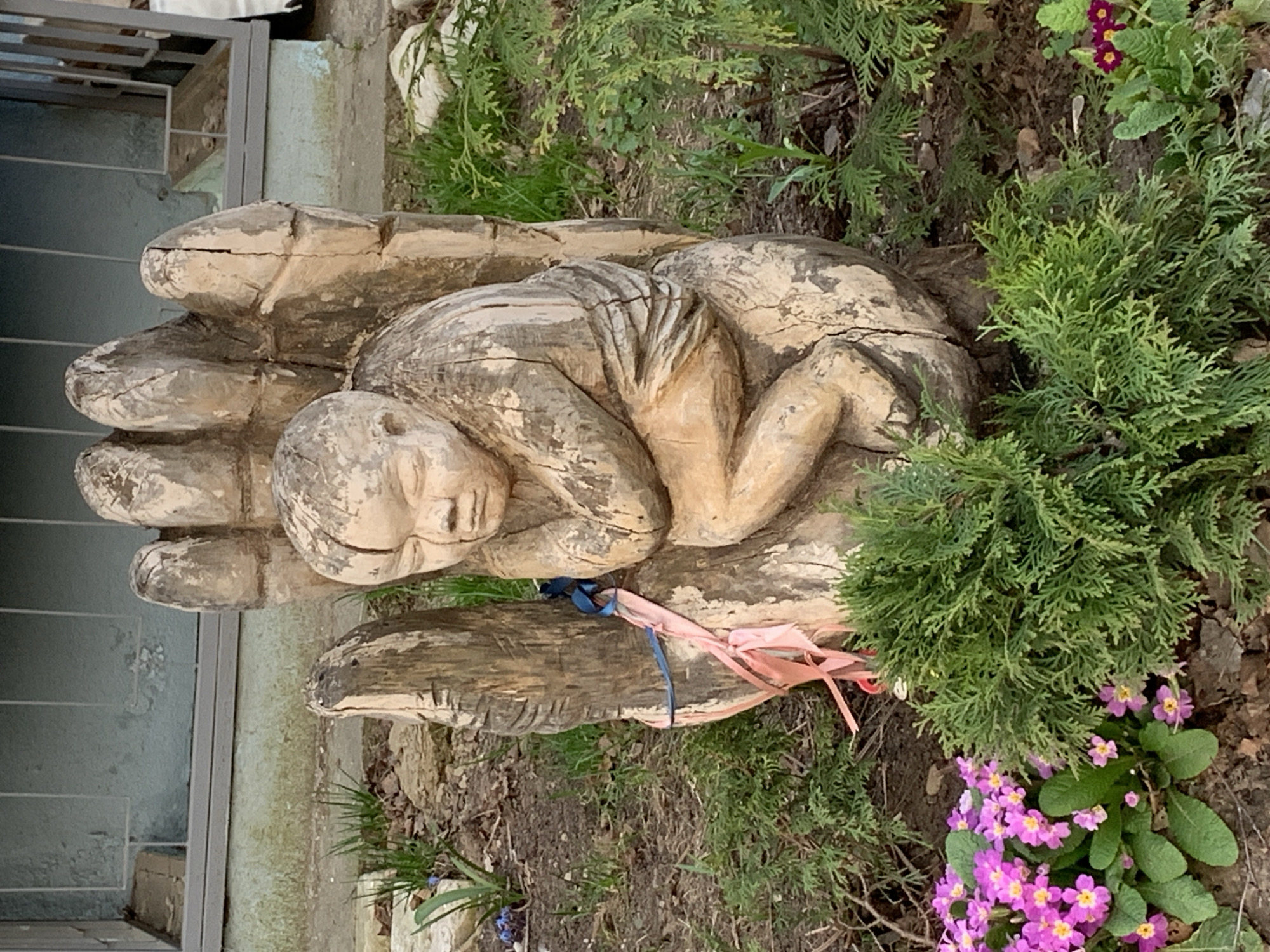 The image depicts a wooden sculpture of a human figure resting in the palm of a hand. The sculpture is weathered, suggesting it is located outdoors. The sculpture is set amongst ground cover vegetation and flowers. Based on the location specified, the image is likely taken outdoors in Chaikovsky, Russia.
