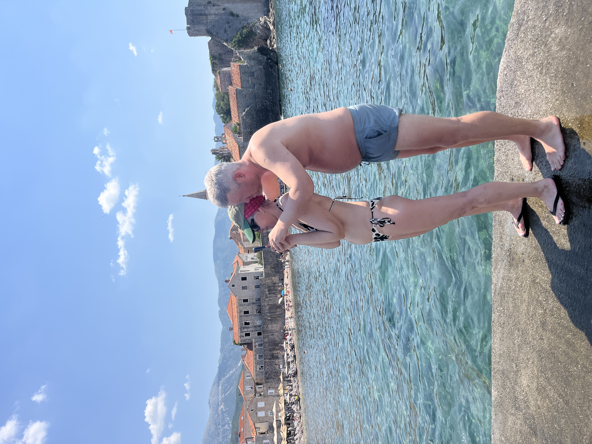 A man and a woman are posing for a photo by the sea in Budva, Montenegro. The man is wearing grey swim shorts and the woman is wearing a patterned bikini. They are standing on a concrete pier with the clear blue water of the Adriatic Sea beside them. In the background, there is a beach with people sunbathing and buildings with red tiled roofs, as well as an ancient stone fortress on a hill. The sky is clear and blue with a few white clouds, suggesting it is a sunny day. The man is leaning in towards the woman, who is holding a phone, as if they are about to kiss or have just shared one. The overall mood appears romantic and relaxed, capturing a vacation moment.