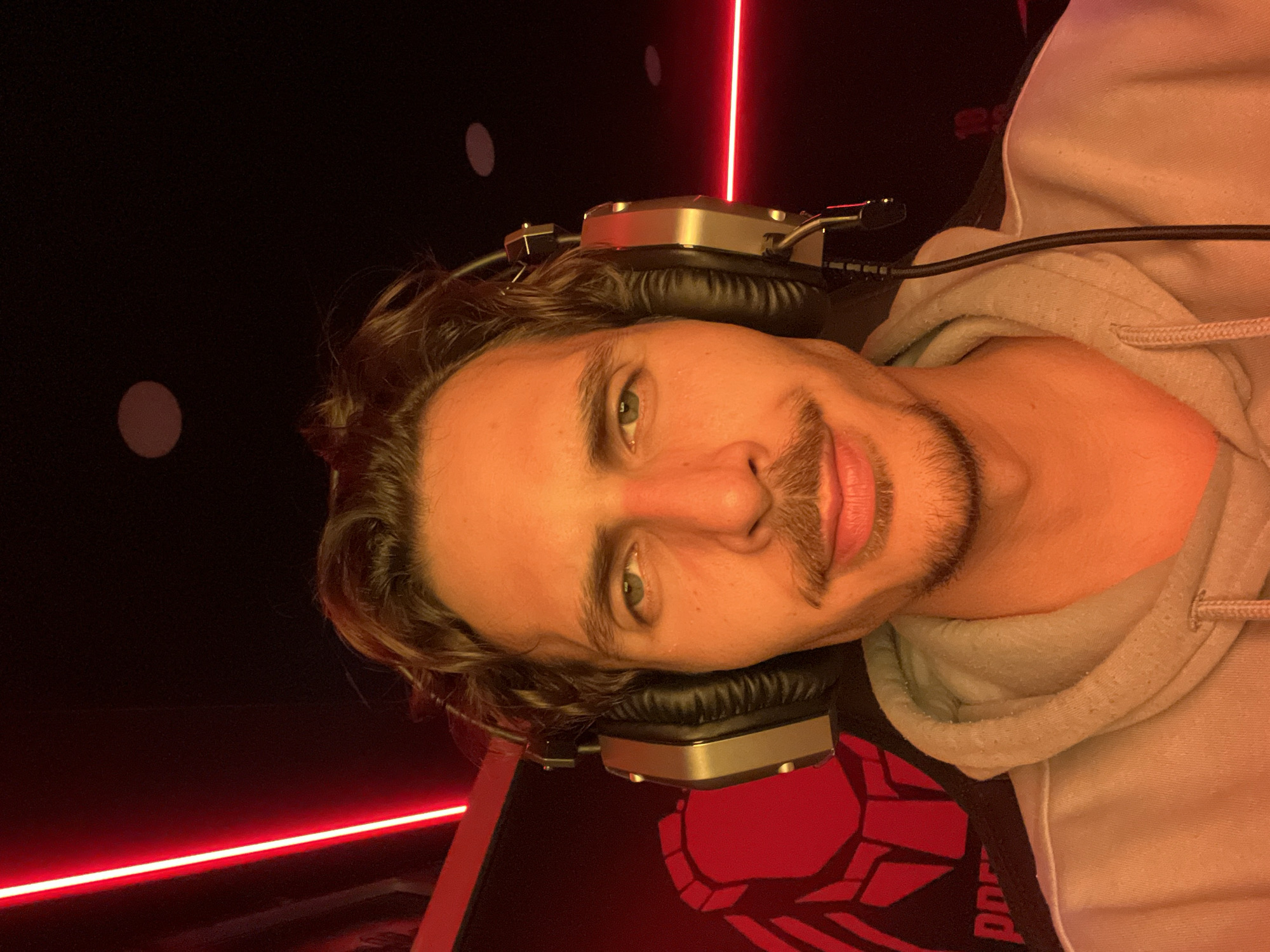 A man with light brown wavy hair, hazel eyes, and a mustache is wearing headphones. He is wearing a light gray hoodie. Behind him is a black wall with white circular lights and red neon lights. A red and black design is visible on what appears to be a surface beneath him. The location in Budva, Montenegro is unknown.
