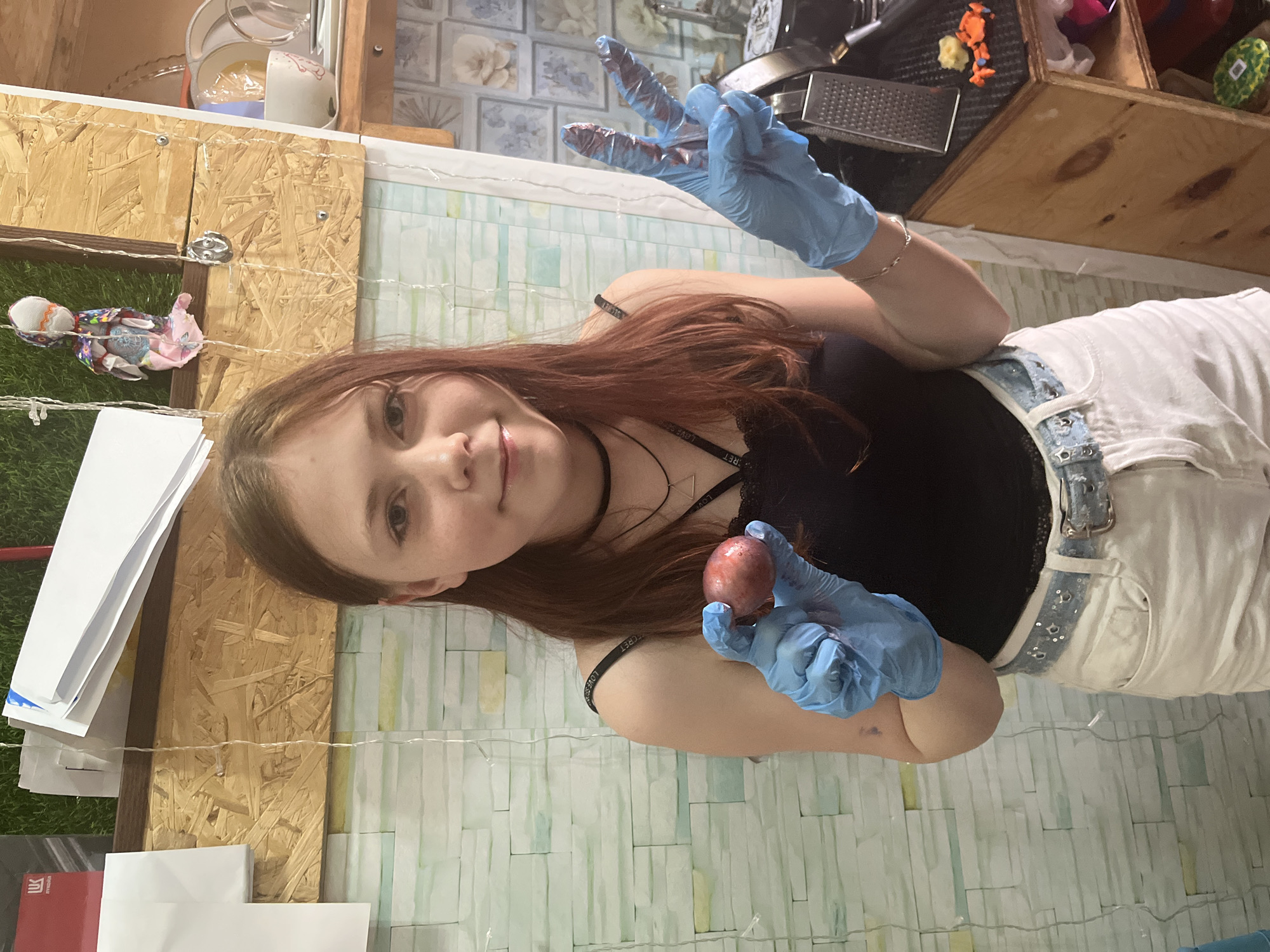 A young woman with reddish-brown hair is posing indoors. She is wearing a black tank top and white denim overalls with a silver studded belt. She is wearing light blue latex gloves. In one gloved hand, she holds a reddish-brown egg, and in the other she is making a peace sign with her fingers. The background consists of a light-colored wall and a shelf with various objects. There is also a wooden structure with artificial grass and papers attached to it. The photo appears to have been taken indoors. No location information is discernible from the image.