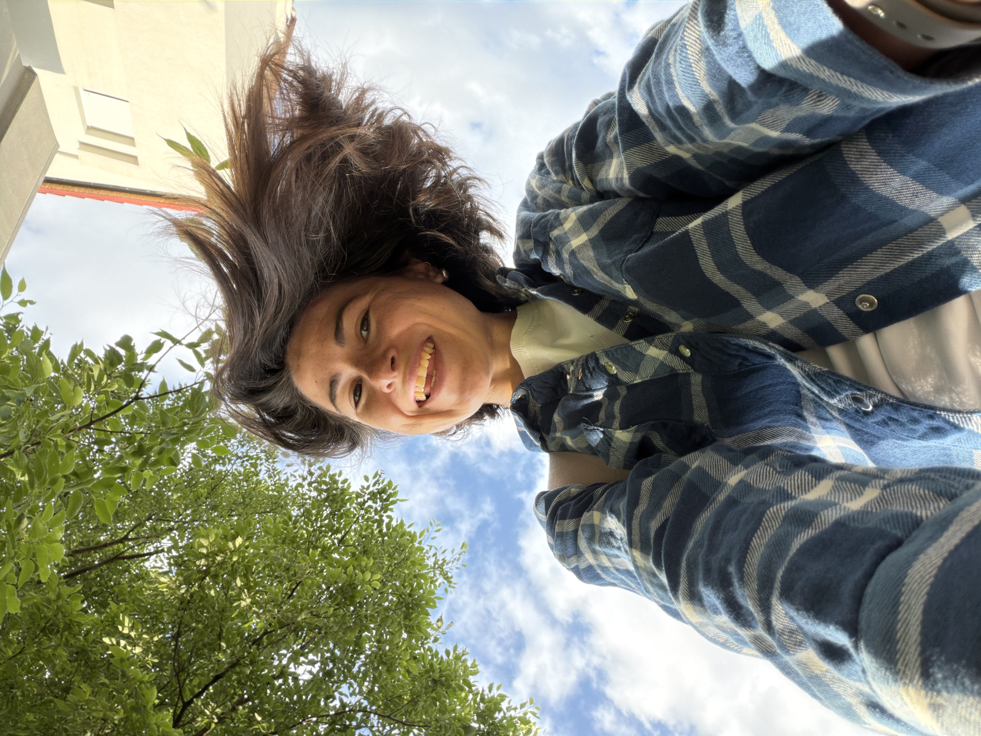 A person is photographed from a low angle, looking directly at the camera with a smile. The person has brown hair and wears a blue plaid shirt over a light-colored shirt. The background consists of green leaves of a tree to the left and the sky with light clouds filling the upper right area of the frame. In the upper left, a portion of a building can be seen. Though not explicitly stated, the image context suggests the photo was taken outdoors, likely in Budva, Montenegro, based on the request. The specific location within the city is not discernible from the image.