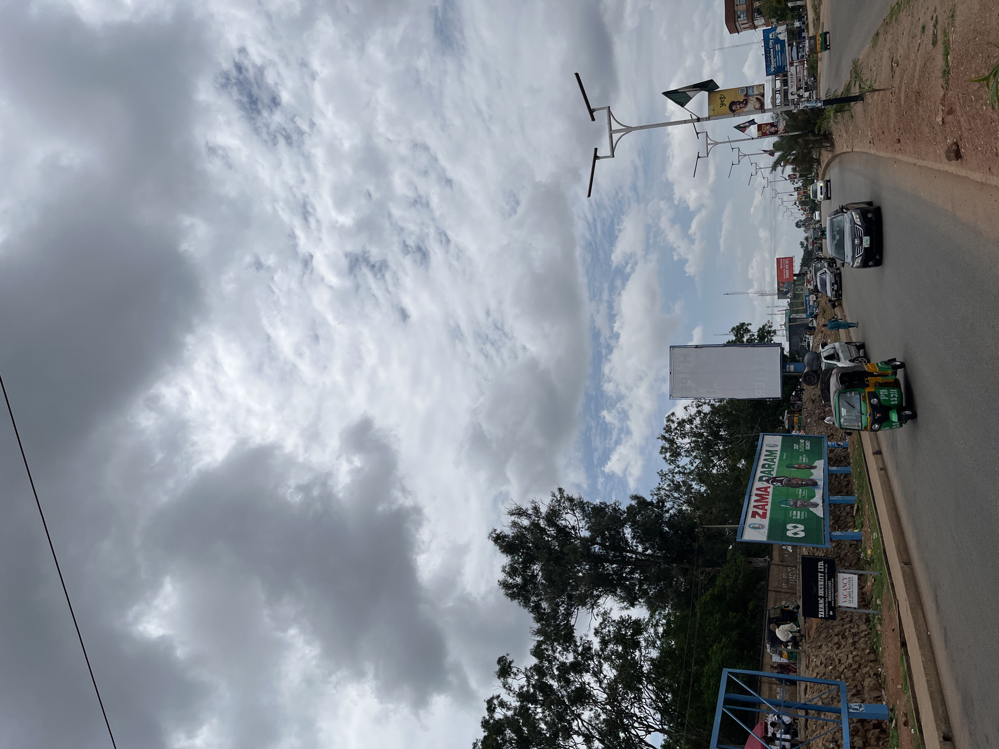 This photograph depicts an urban street scene in Jos, Nigeria, under an overcast sky with patches of blue. The upper portion of the frame is dominated by heavy grey clouds, while lower, lighter clouds reveal glimpses of blue sky towards the horizon on the right. A dark power line crosses the very top of the image.
The foreground features a multi-lane asphalt road curving gently to the right in the distance. A green and yellow tricycle (keke Napep) with the identifier "PTA 1135I" is prominent on the left side of the road, moving away from the viewer. A black sedan (Toyota Camry) travels towards the viewer in the center lane. Other vehicles, including cars and tricycles, are visible further down the road, indicating traffic. A person in a light blue tunic walks on the left side of the road, parallel to the black sedan.
On the left side of the road, a large blue metal frame stands in the immediate foreground, partially obscuring an unpaved area. Behind this, an earthen embankment rises, dotted with rocks and sparse vegetation. A prominent billboard in green and white displays "ZAMA DARAM O" alongside images of three male political figures and the text "2027 PLATFORM NO VACANCY," suggesting an election campaign. Below it, a smaller black sign for "TARMAC SECURITY LTD." advertises "VACANCY GUARDS WANTED." Behind these structures, dense green trees and shrubs are visible, along with glimpses of people and activity further back on the roadside.
The right side of the road features multiple tall street light poles equipped with solar panels and horizontal arms, extending into the distance. A green flag, possibly a national or political flag, hangs from one of these poles. Another billboard displays "Yes!" and an image of a person, while further down, a "DOMINION CITY" billboard is visible. The roadside here consists of a brown dirt shoulder with scattered small plants and sparse trees, leading to buildings in the background.