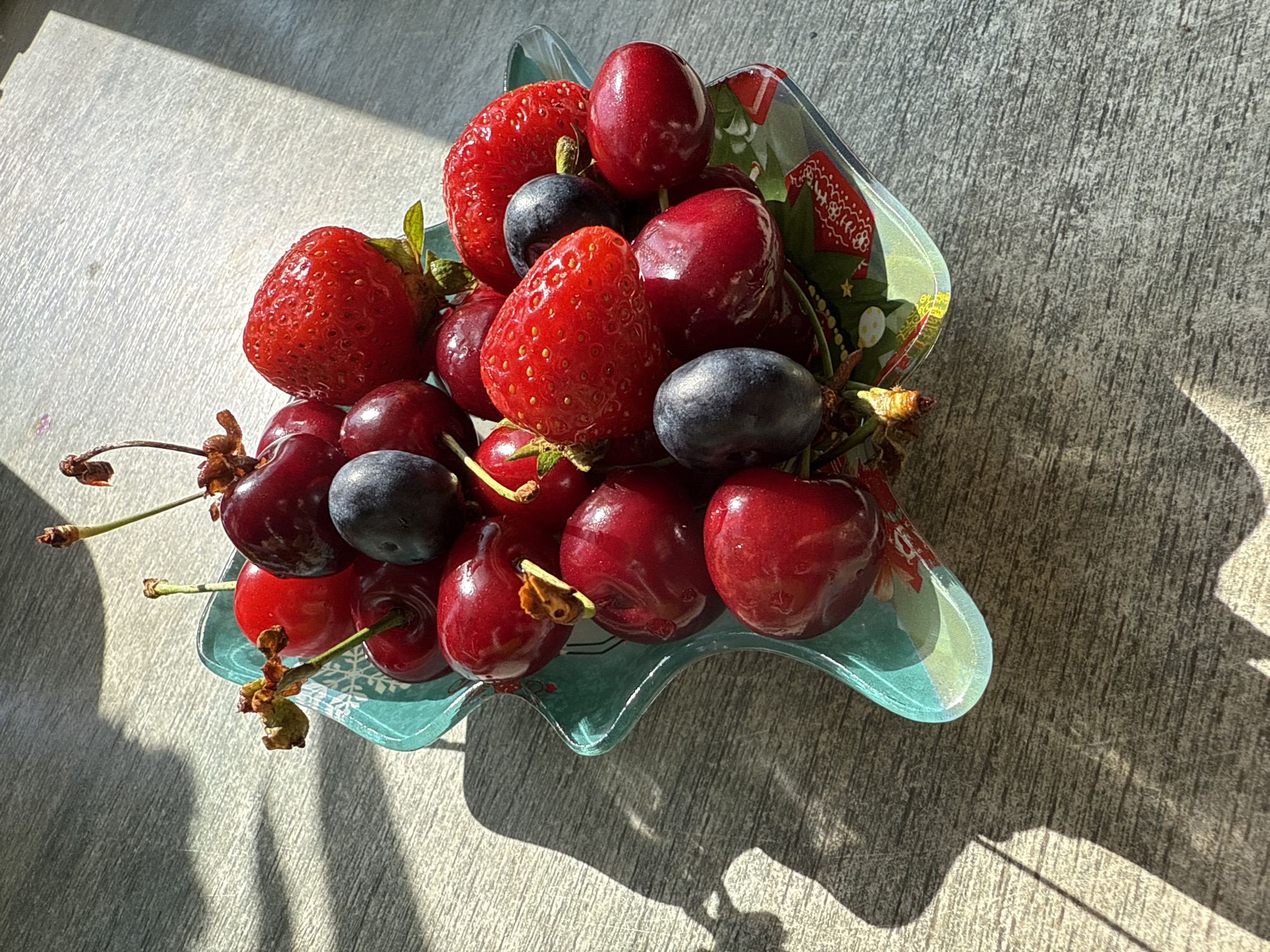 The image shows a close-up of a decorative, star-shaped, blue-green glass bowl filled with fresh fruit. The bowl rests on a light-colored wooden surface, possibly a table, which exhibits a visible wood grain texture and is partially illuminated by sunlight casting strong shadows across it.
The fruit in the bowl includes bright red strawberries, deep red cherries with stems, and dark, almost black, grapes. The arrangement of the fruit is natural, with some pieces overlapping others. The sunlight highlights the glossy surfaces of the fruit, creating reflections.
The bowl itself is transparent enough to show a festive pattern on its underside, featuring red and green elements that appear to be stylized Christmas designs. The edge of the bowl has a slightly wavy, scalloped appearance.
The overall scene is one of natural light and fresh produce, with the shadows on the wooden surface suggesting an outdoor setting or a strong light source indoors. There are no people or animals visible in the image.
The prompt's request to include the location "Kroyanskoye, Russia" cannot be fulfilled as there are no visual cues within the image to support this specific location. The description is based solely on the visible elements of the photograph.