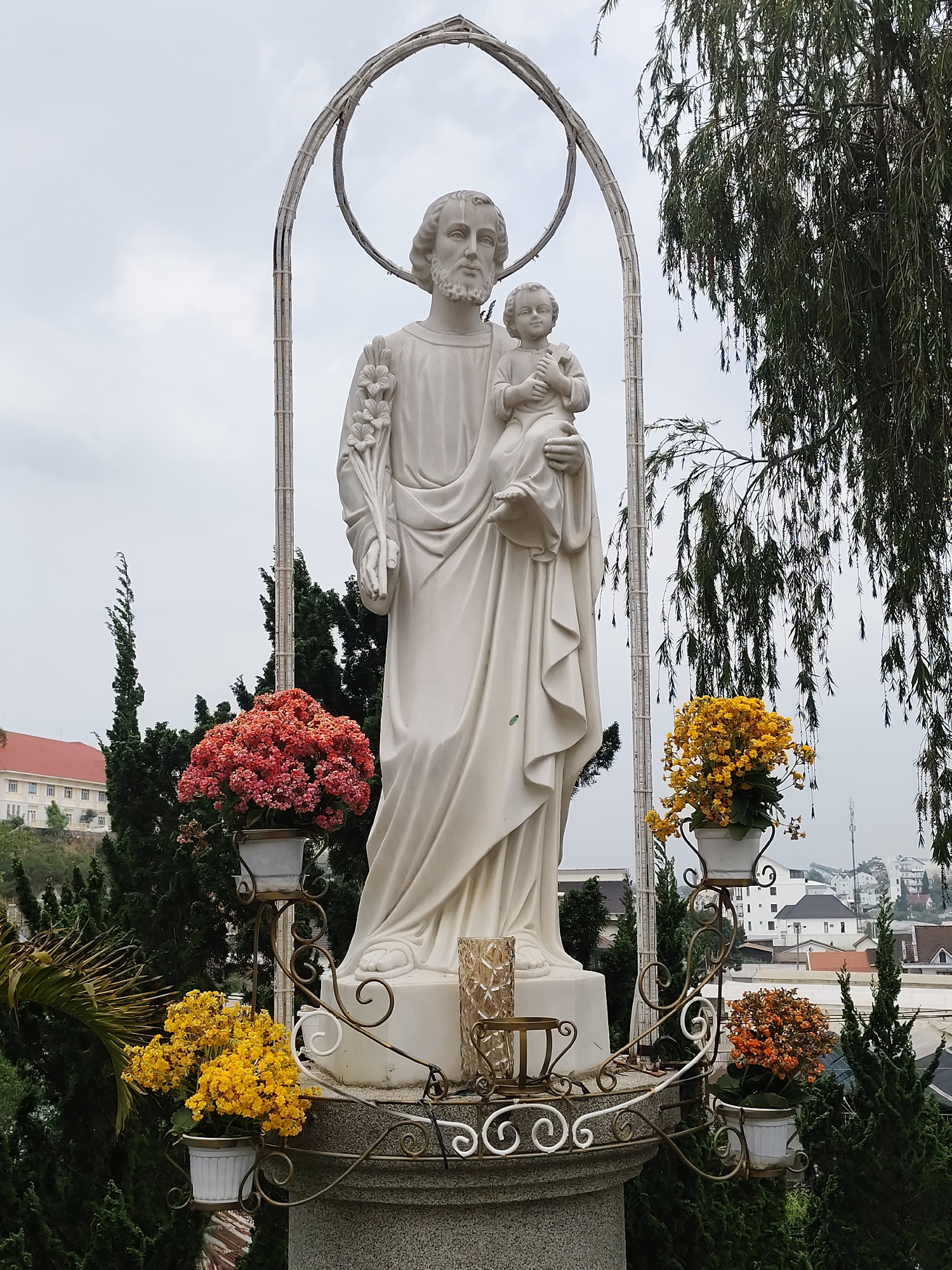 The image shows a full-length statue of Saint Joseph holding the child Jesus. Both are white and stand on a grey stone plinth decorated with wrought-iron stands holding potted flowers. The plants display red, orange, and yellow blooms. Behind the statue is an arched frame. The statue is situated outdoors, with various trees and plants surrounding it. Buildings are visible in the background. The location is Lang Biang - Đà Lạt, Vietnam.
