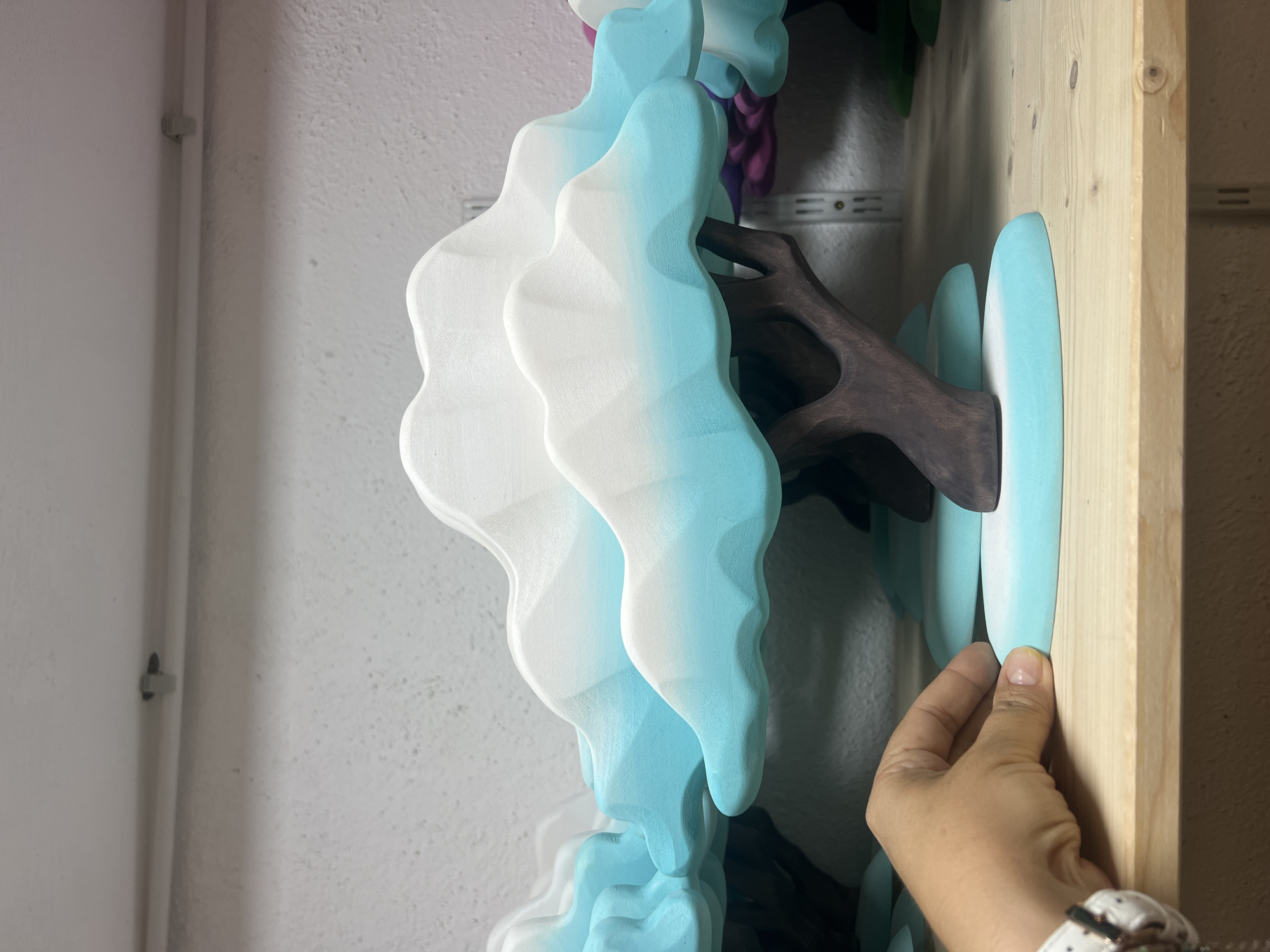 A person's hand is holding the base of a wooden, stylized tree sculpture. The tree has a dark brown trunk and branches that support a cloud-like canopy. The canopy is painted with a gradient of white at the top transitioning to a bright turquoise color towards the bottom. The sculpture sits on a light blue oval base, which rests on a wooden shelf. Behind the sculpture, more similar-looking sculptures are partially visible, with one having a purple element. The background appears to be a white textured wall with a white pipe running horizontally across the top, secured by gray clips. The lighting is even and appears to be artificial. The scene is indoors, likely in a workshop or display area.