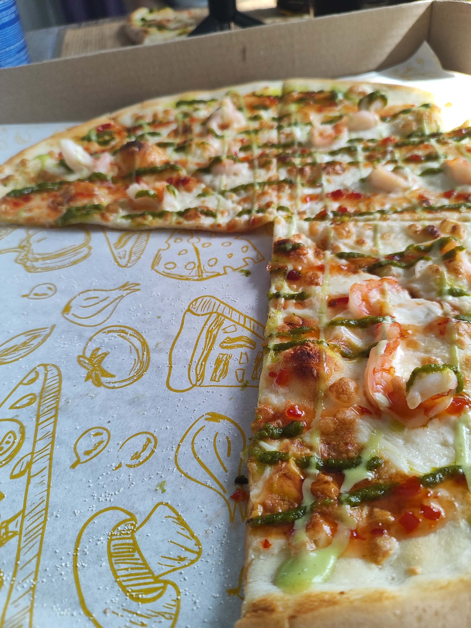 The image displays a high-angle, close-up view of a partially consumed rectangular pizza situated within an open brown cardboard pizza box. The pizza is topped with a white cheese base, pinkish-orange shrimp, a vibrant green pesto-like sauce drizzled in parallel lines, and scattered red droplets of another sauce. It has been pre-cut into rectangular slices, with several pieces already removed from the left side. The white parchment paper lining the pizza box is visible beneath the missing slices and features yellow outline illustrations of various food items, including cheese, tomatoes, and onions. Numerous small, white granular particles, consistent with cornmeal or flour, are scattered across this paper liner, particularly on the left. In the blurred background, a segment of another pizza or remaining slices is visible within a separate box, alongside a blue cylindrical object on the far left and an indistinct dark object on the upper right. The scene is illuminated by natural light originating from the right side. No individuals are depicted in the image. This scene is located in Marusino, Russia.