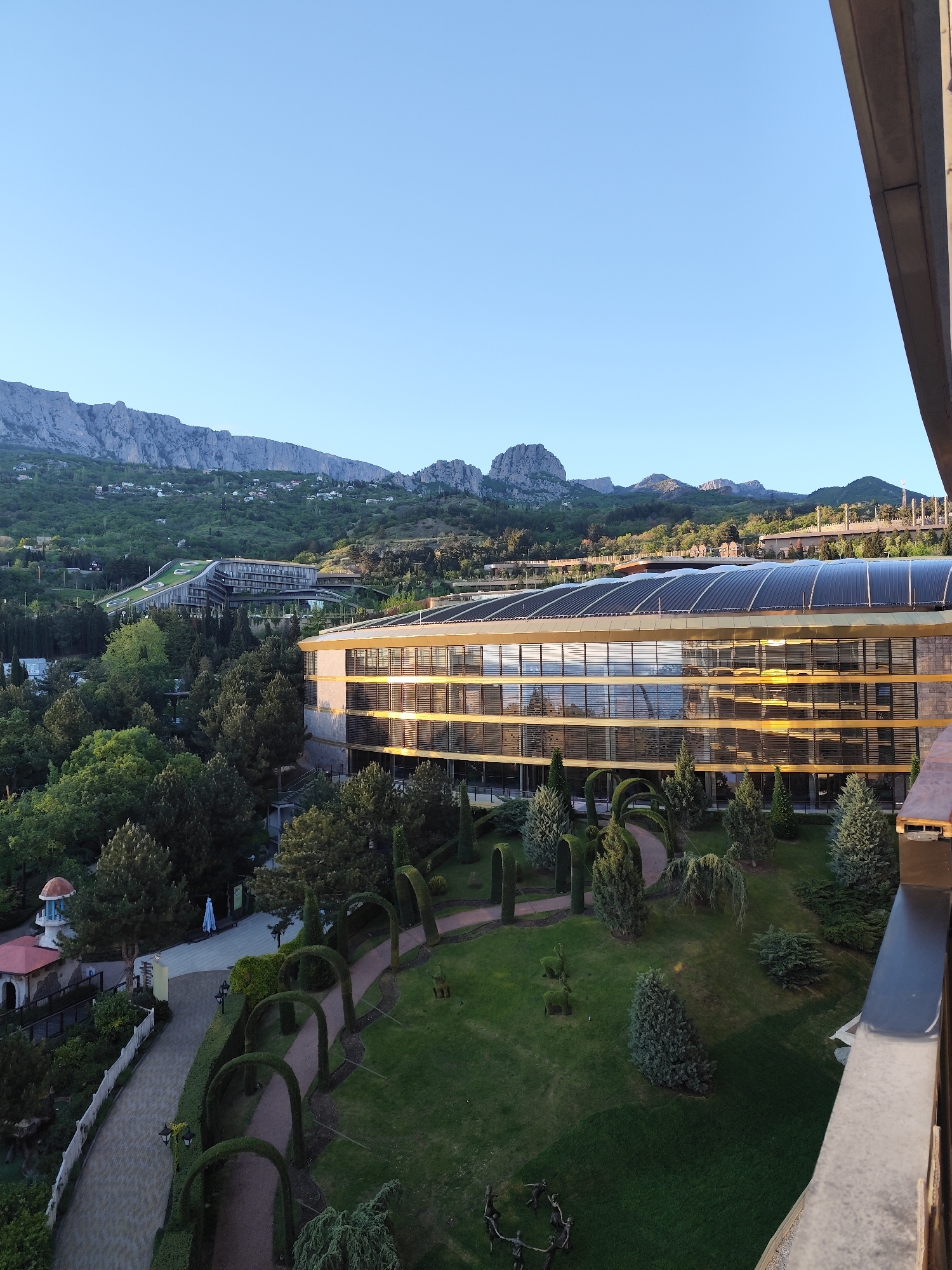 The image shows a daytime view from a high vantage point, possibly a balcony, overlooking a landscaped area in Симеизский поселковый совет, Ukraine. The scene is dominated by a modern, multi-story building with a curved roof and a reflective glass facade, suggesting it may be a hotel or resort. The building is surrounded by meticulously maintained gardens featuring arched hedges, neatly trimmed trees, and winding pathways. In the distance, a mountain range provides a scenic backdrop. The sky is clear and blue, indicating fair weather. The image captures a sense of tranquility and showcases the blend of modern architecture with natural surroundings.
