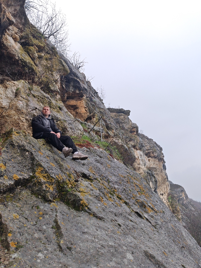 An adult male is depicted seated on a grey, lichen-covered rock slope, part of a larger cliff formation in the Shumen Plateau Natural Park, Shumen, Bulgaria. The male, with short light-colored hair, is wearing a dark puffer jacket over a grey hooded sweatshirt, black pants, and light brown hiking shoes. He is positioned facing slightly right of the camera, looking directly forward with a neutral expression. His legs are extended downwards along the incline of the rock, hands resting on his knees. The rock face behind and above him exhibits layers of beige, brown, and grey stone with sparse, bare deciduous vegetation growing from crevices. The sky is uniformly overcast and light grey, suggesting a cloudy day. The overall environment is rugged and natural.
