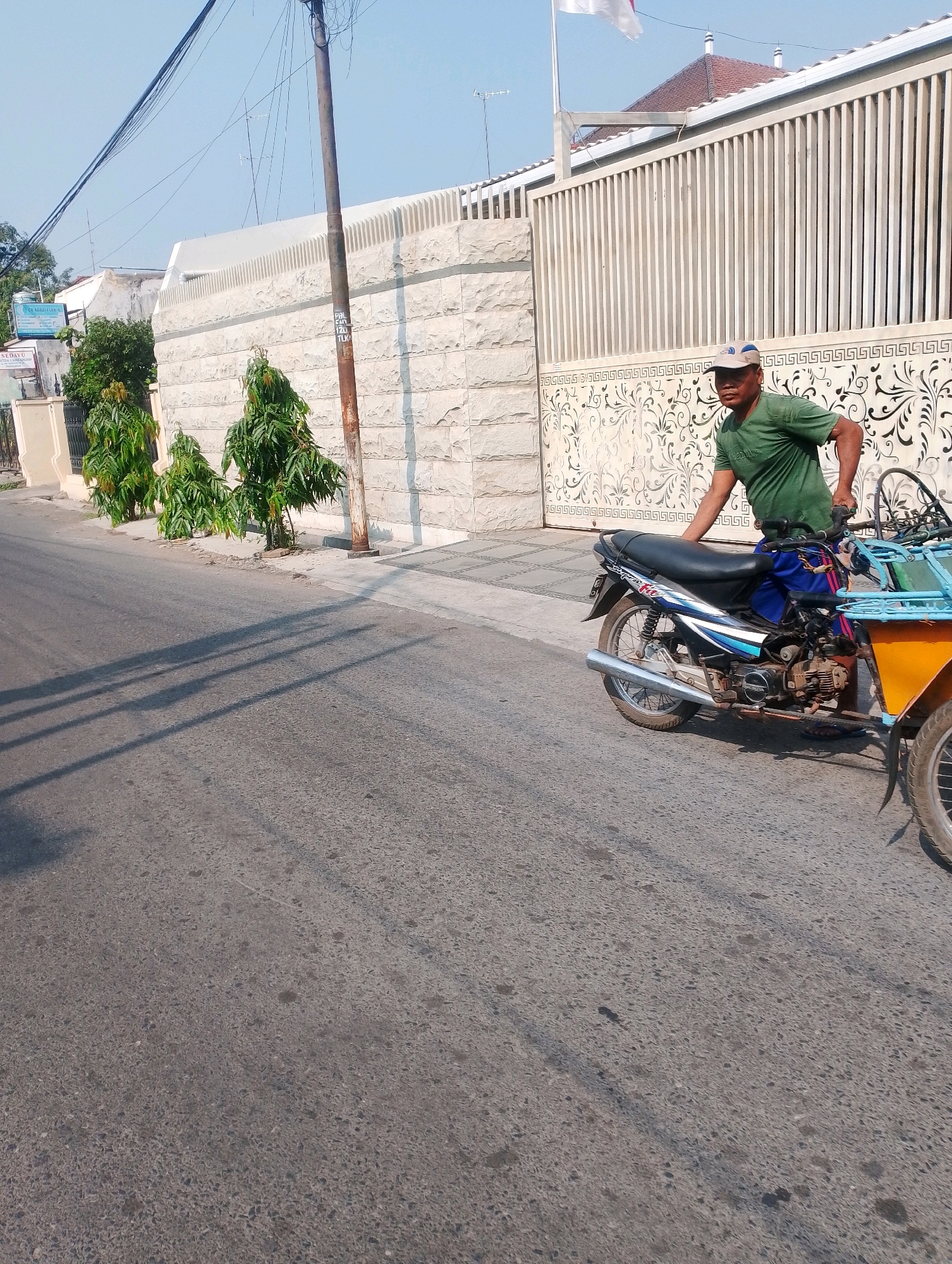 A man wearing a green t-shirt and a blue baseball cap is standing next to a motorcycle with a sidecar on a paved road in Probolinggo, Indonesia. The motorcycle is blue and black and has a silver exhaust pipe. The sidecar is yellow and has a blue frame. The man is holding onto the motorcycle with his right hand. In the background, there is a white stone wall with a decorative pattern and a tall metal fence. There are also some green trees and a utility pole with wires. The sky is clear and blue. The sun is shining, casting shadows on the road. The scene appears to be in a residential or commercial area.