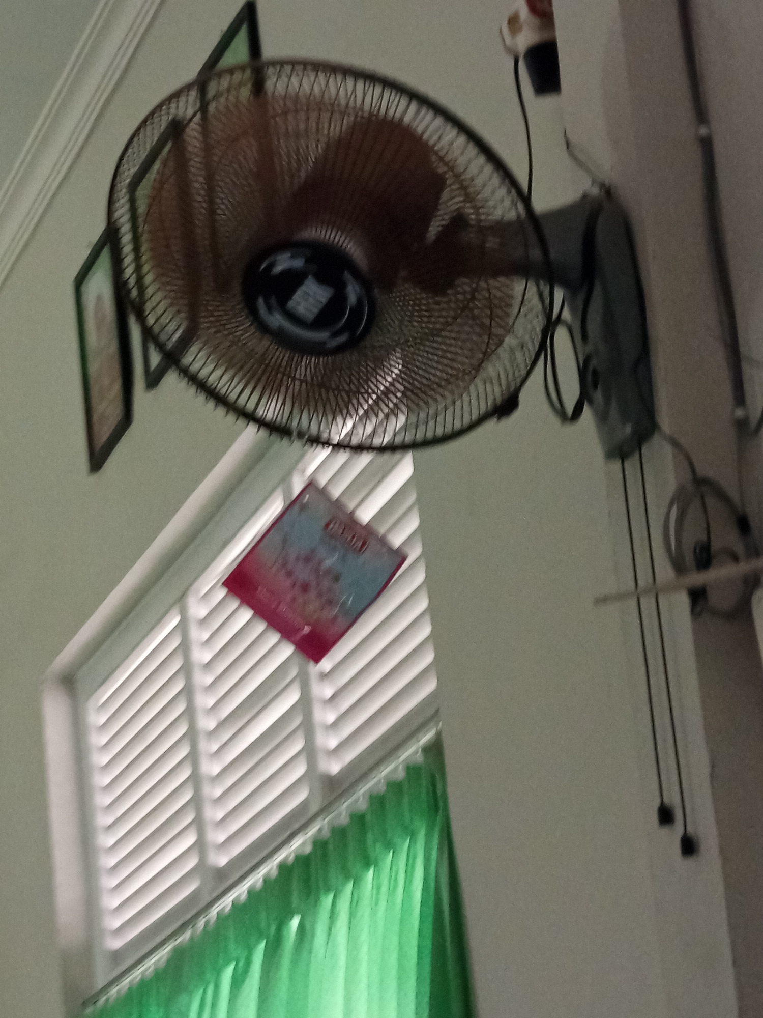 The image shows an indoor scene, likely a room, with a wall-mounted fan prominently displayed in the upper portion. The fan has a dark frame and a brown oscillating head. Below the fan, a window with horizontal white blinds is visible, partially covered by bright green curtains. A small, rectangular object, possibly a book or a folder, with pink and blue colors and some writing or images on it, is hanging from the fan's guard. Two framed pictures are mounted on the wall to the left of the fan, one appearing to be larger than the other. The lighting is dim, casting shadows and creating a somewhat muted atmosphere. The setting is an interior space in Lumutan, Indonesia.