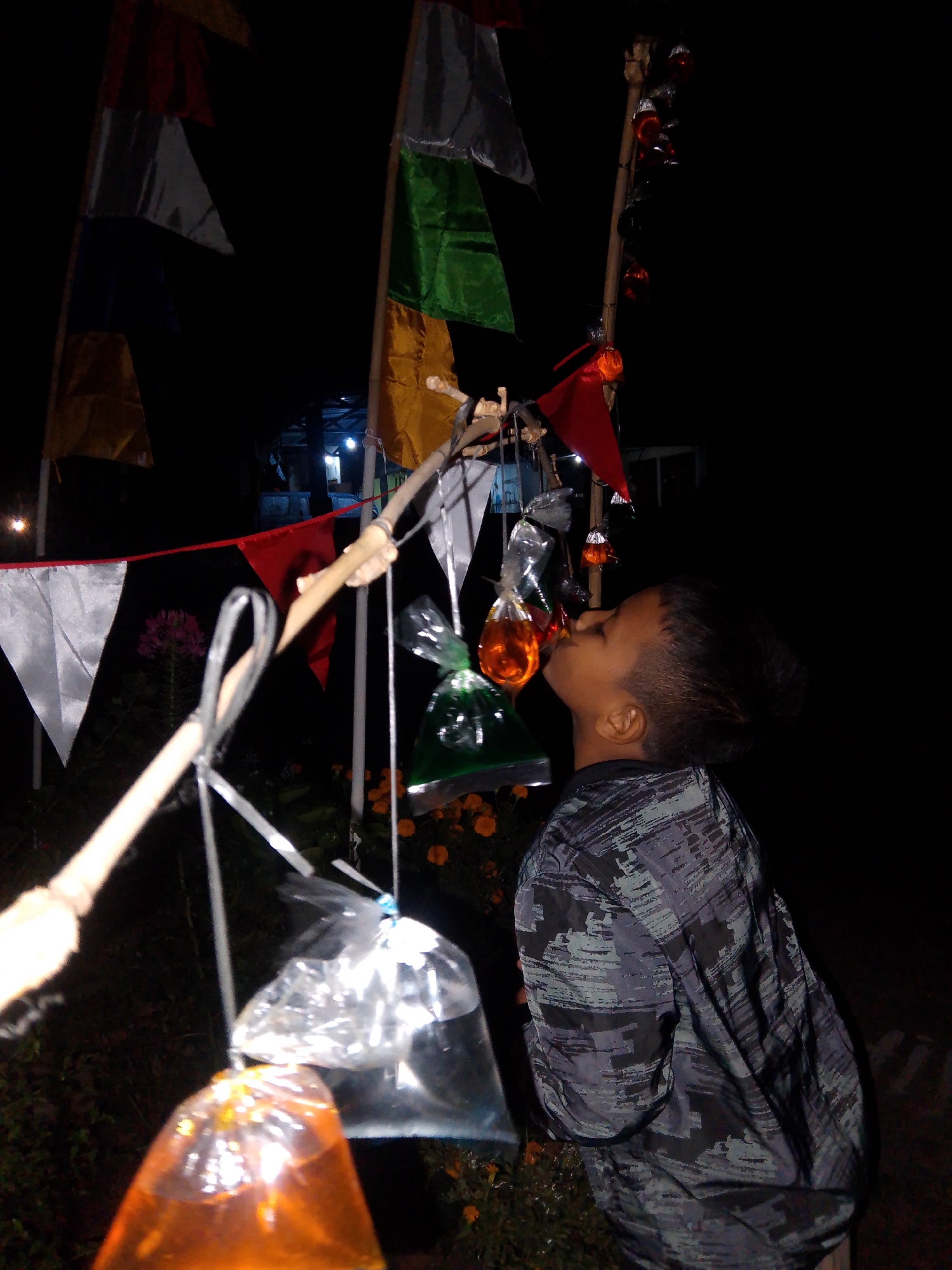 A young boy is being filmed at night near the city of Bantar, Indonesia. The boy is standing in front of several flags and is trying to drink from a bag filled with orange liquid. The flags are multi-colored and appear to be made of fabric. Some of the flags are red, white, green, and yellow. The boy is wearing a patterned shirt and has his mouth open as he attempts to drink from the bag. The background is dark and appears to be a natural setting with some foliage. There are also some lights visible in the distance.