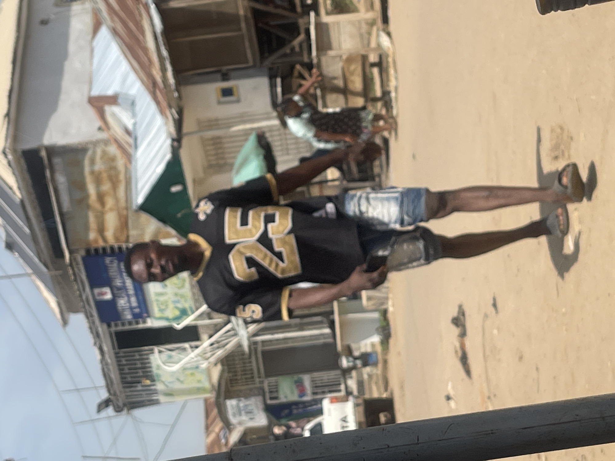 A man in a black and gold jersey with the number 25 walks down a dusty street in Abuja, Nigeria. He is wearing denim shorts and sandals. Behind him, buildings with corrugated metal roofs line the street. A white pickup truck is parked on the left side of the road. In the background, a woman in a light blue shirt and dark patterned skirt leans over a table. The overall scene depicts a typical streetscape in a bustling Nigerian city.