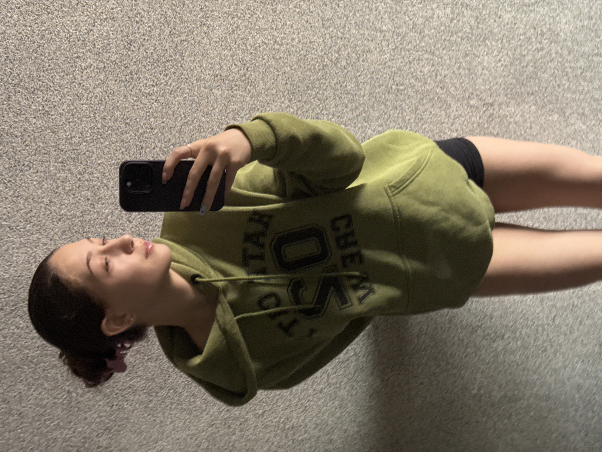 A young woman is captured in a mirror selfie, seen in profile, holding a black smartphone with her right hand. She is wearing a loose-fitting, olive-green hooded sweatshirt. On the front of the hoodie, dark, blocky letters are partially visible, appearing to spell out "TALK OVER CREW" or a similar phrase. Underneath the hoodie, black shorts are visible. She wears a small ring on her right ring finger, and her nails are painted with a light blue and white pattern. Her dark hair is pulled back into a bun or messy updo, secured with a small, light purple hair clip.
Her eyes appear closed or are cast downwards, giving her a serene or neutral expression. Her lips have a slight sheen. The background is a uniform, light gray, textured surface, possibly a carpeted floor or a textured wall, indicating an indoor setting. The lighting is bright and even, suggesting daytime. The overall composition is casual, capturing a moment as she poses for or takes the photo in what appears to be her reflection.