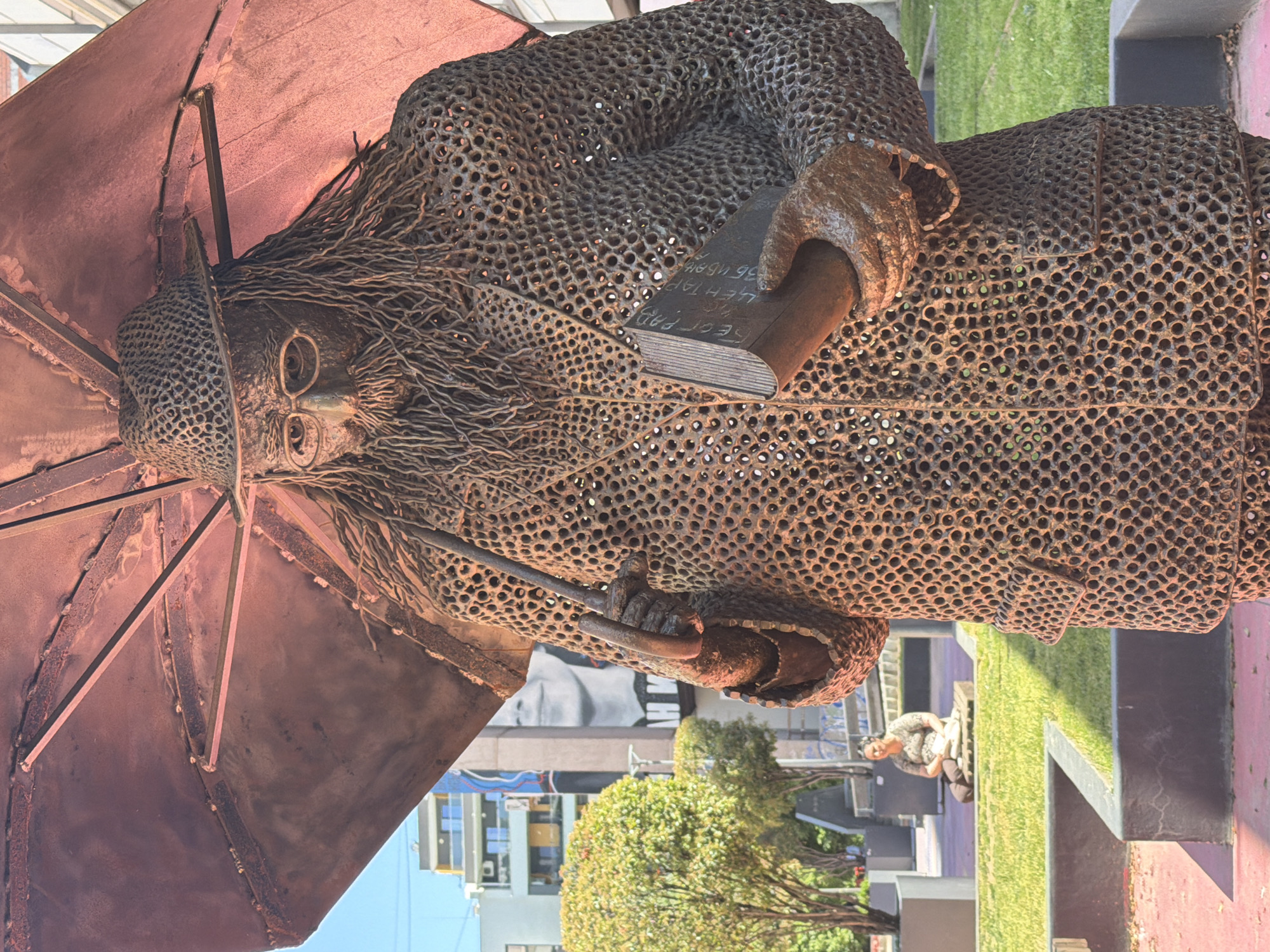 The image depicts a sculpture of a man holding an umbrella and a book, constructed from a metal mesh with circular perforations. The sculpture is located in Palilula Urban Municipality, Serbia. The man is wearing a hat and coat, and the umbrella provides partial coverage over his head. The book has visible text on its spine. The sculpture is situated in a park or public space with grass, trees, and other greenery visible in the background. Other buildings are in the distance. A person is visible sitting on a bench nearby. The sculpture appears to be made of aged or rusted metal.