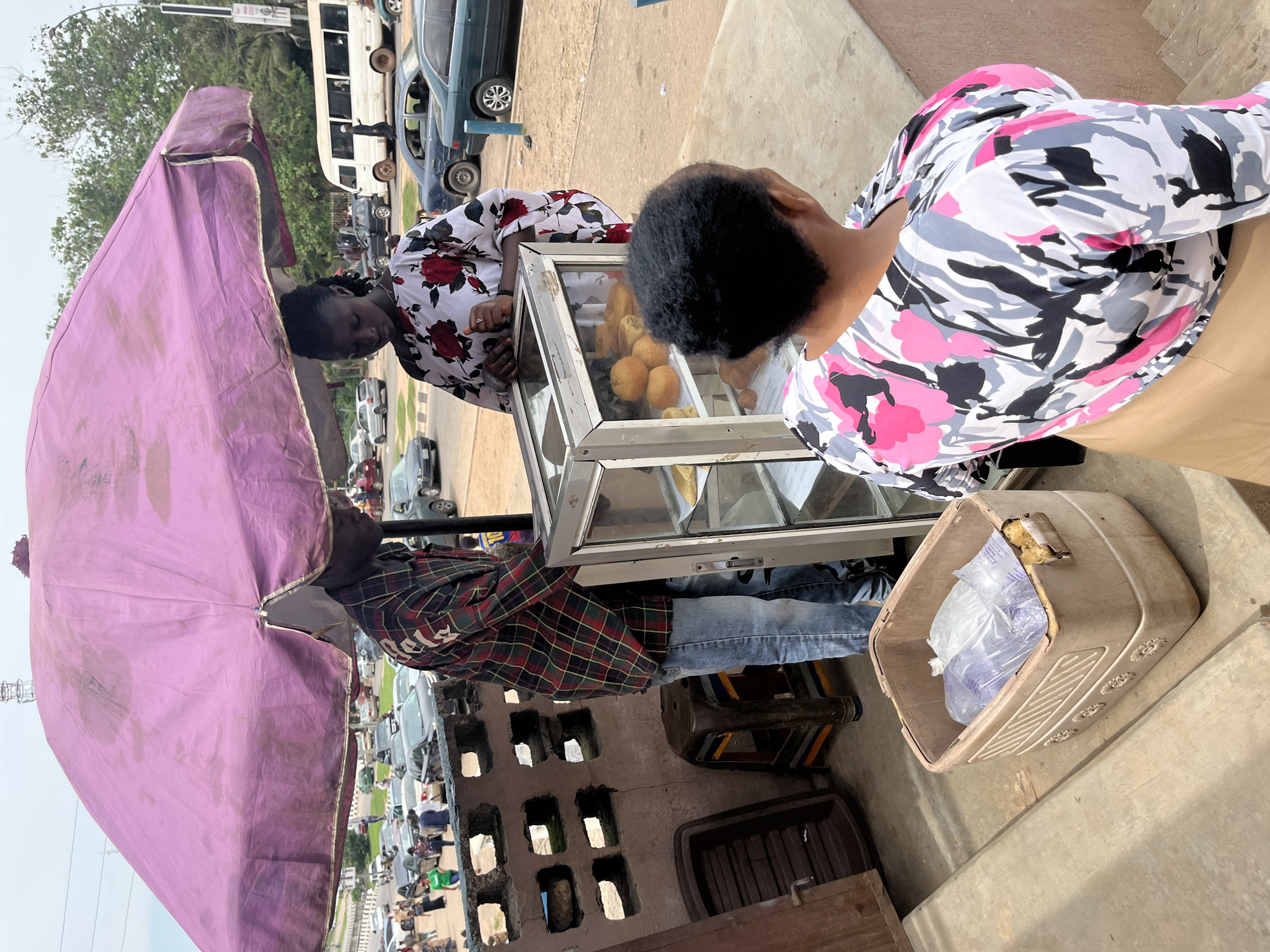 In Abuja, Nigeria, under a large, faded purple umbrella, a street vendor displays baked goods in a glass-fronted case. The vendor, a young man wearing a plaid shirt and jeans, stands to the left of the case. A young woman in a floral dress stands opposite him. Behind them, a woman with dark, curly hair, wearing a colorful, abstract-patterned shirt, faces the display case, presumably as a customer. In the background, traffic on a busy street is visible, including cars, a white bus, and pedestrians. The scene is set on a paved surface, with concrete steps and a textured wall to the left of the vendor. A plastic container filled with plastic-wrapped items sits on the steps.