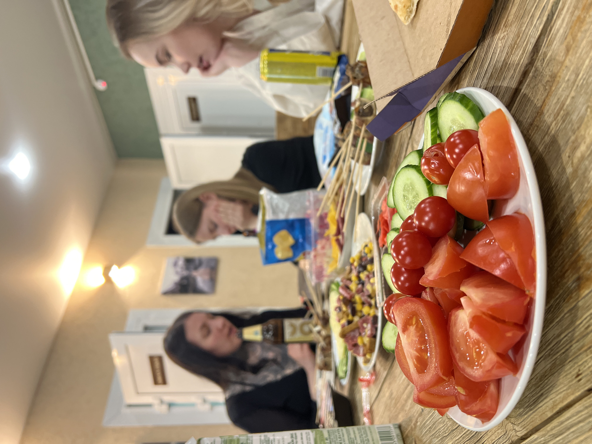 A photograph, likely taken indoors, shows three women around a table laden with food, seemingly enjoying a meal together.
The foreground is dominated by a white plate filled with sliced tomatoes and cucumbers. Moving back, the table holds skewers, salads, and what appears to be pizza in a cardboard box. In the background, three women are visible. One, with blonde hair, is facing down. Another, with brunette hair, is covering her face with her hand. A third woman with dark hair is drinking.
The scene is set indoors, with walls painted in light colors. Pictures hang on the walls in the background. Based on the location information provided, the scene is likely taking place in Chaikovsky, Russia. The image suggests a casual gathering, perhaps a meal amongst friends.