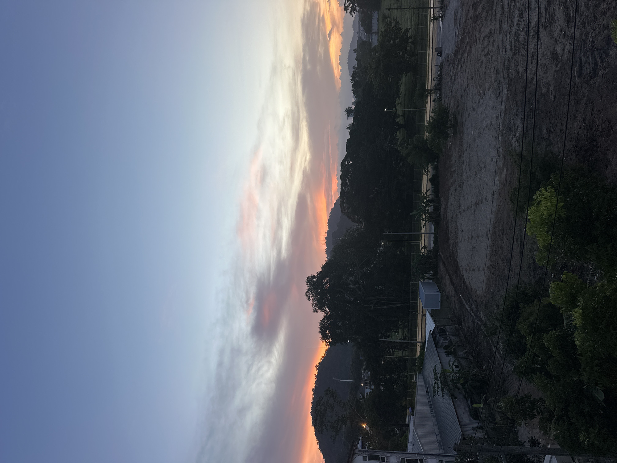 This image depicts a sunset over a semi-urban area, possibly in or near Wichit, Thailand, characterized by a gradient sky transitioning from pale blue at the top to hues of orange and pink near the horizon. A chain of mountains forms the background, with several tall, slender structures, possibly communication towers or lampposts, visible on their slopes and summits. Dense foliage, comprised of large trees and smaller bushes, dominates the middle ground, obscuring much of the low-lying structures. To the left, a portion of a white building with visible windows is present. In the foreground, a cleared, dirt lot is visible, marked by tire tracks and some scattered debris, suggesting recent activity or preparation for construction. Electrical wires run horizontally across the lower portion of the frame, intersecting with the foliage. The lighting indicates it is either early morning or late evening, with the sun low in the sky, casting a soft, diffused light. There are no visible people in the scene.