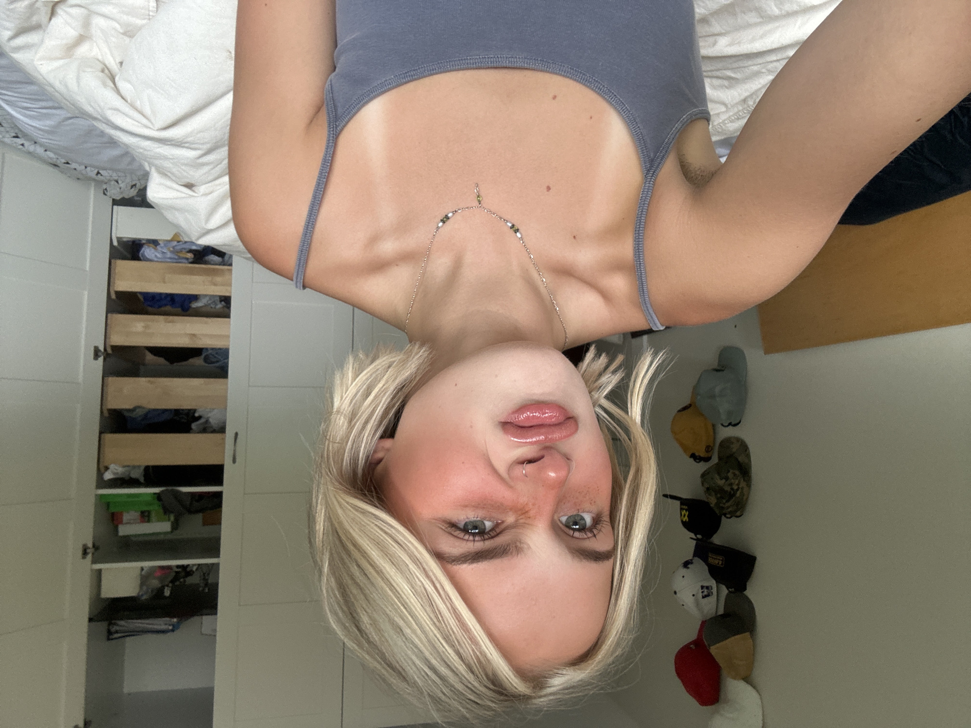 The media file is a portrait-oriented selfie taken indoors. The main subject is a young person with blonde hair, who is facing the camera with their head tilted upside down. They are wearing a grey tank top and a delicate necklace. Their facial expression is playful, with bright eyes and slightly parted lips. The background shows an open closet with shelves filled with clothing and various items, as well as a wall with a row of baseball caps. The lighting suggests it is daytime. The presence of a closet and the caps indicates a casual, domestic setting. There is no visible text in the image.