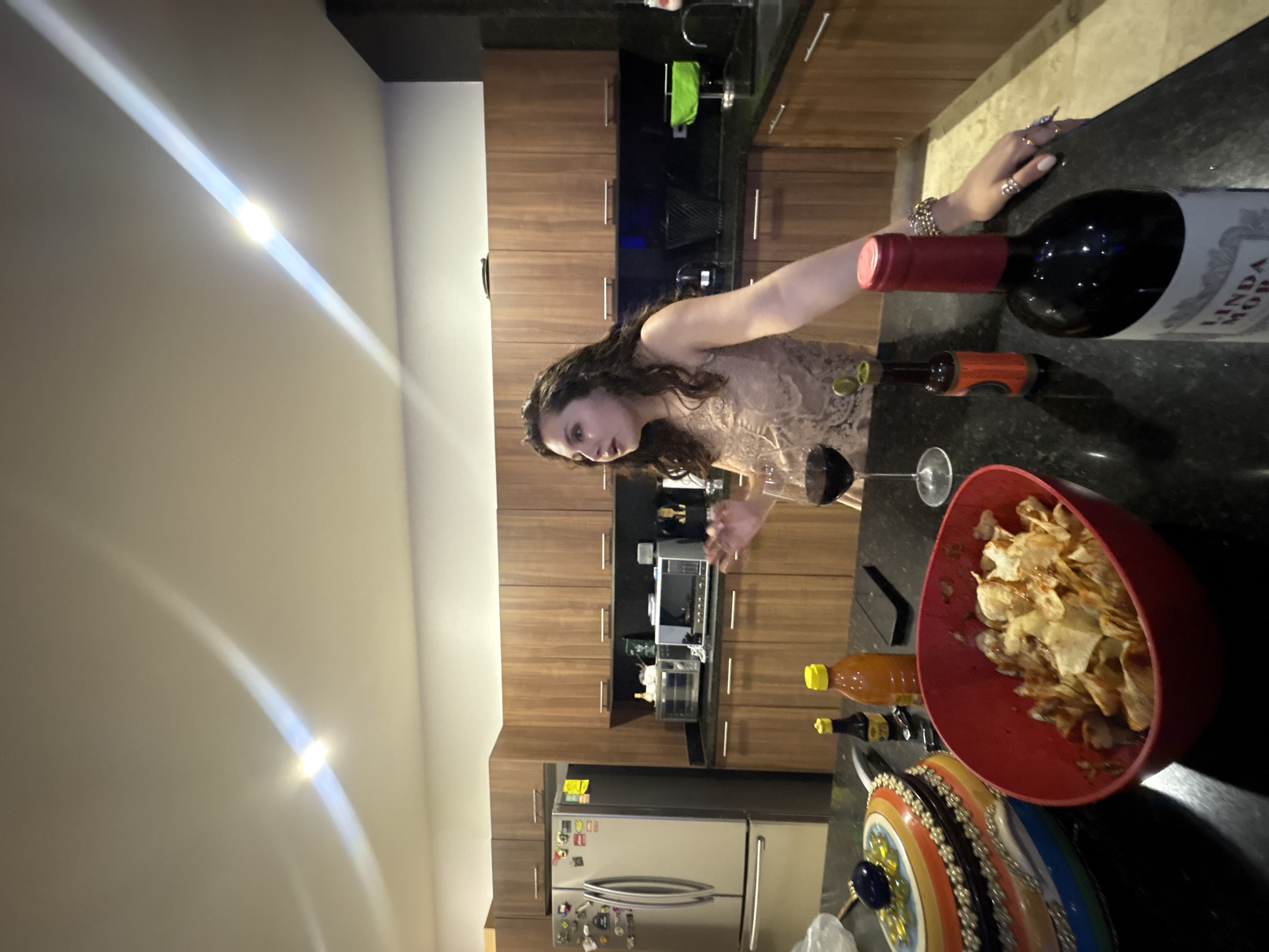 A woman in a beige dress is standing in a kitchen. She is next to a counter, on which there is a bottle of Linda Mora wine, a glass of wine, a red bowl of chips, and two bottles of hot sauce. Behind the woman is a kitchen with dark wood cabinets and a stainless steel refrigerator. The kitchen is in a home in Santa Clara Ocoyucan, Mexico.