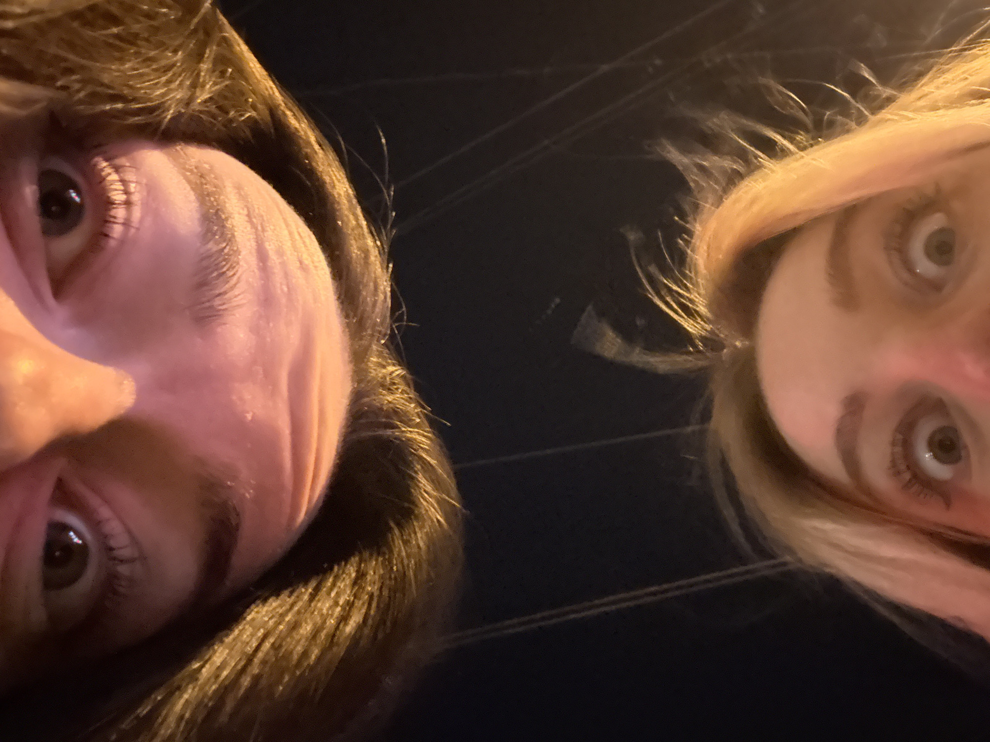 Two individuals are captured from a low angle, emphasizing their faces. The individual on the left has dark hair, while the individual on the right has blonde hair. Both individuals are looking directly at the camera with wide eyes. The background appears dark, with faint lines suggesting overhead wires or cables. The photograph appears to have been taken outdoors at night, with minimal ambient light. The location cannot be identified as Krasnoyarsk, Russia, from the visual information provided.