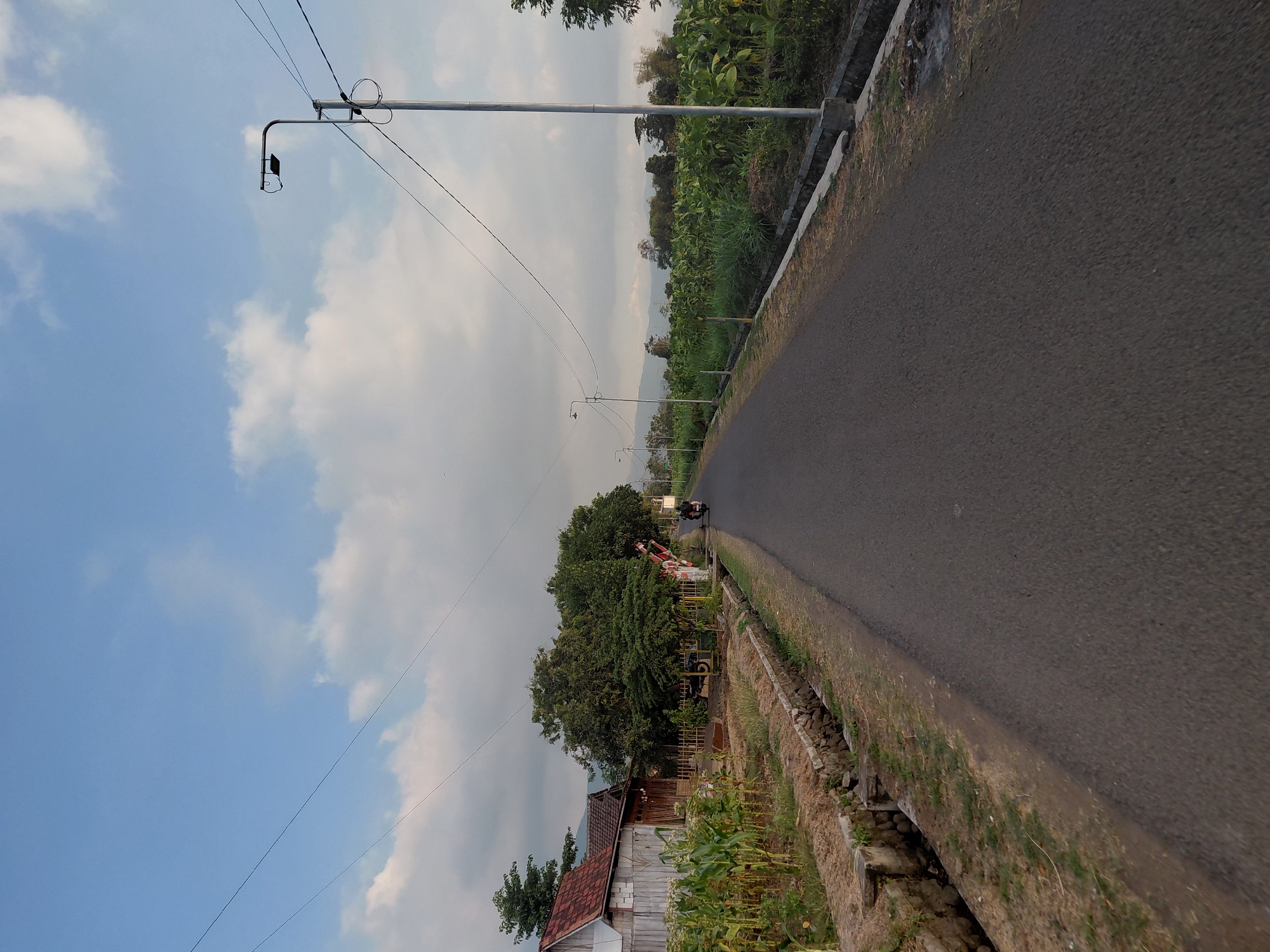 The image depicts a rural road in Kedungrejoso, Indonesia, under a partly cloudy sky. The asphalt road stretches into the distance, flanked by vegetation and agricultural fields. On the left side of the road, a small wooden house with a red tiled roof is visible, adjacent to a cornfield and a drainage ditch. Further down the road, a few trees and a red and white barrier suggest a crossing, possibly for a railway or a local entrance. A person on a motorcycle is traveling away from the camera on the right side of the road. Several lampposts line the road, with electrical wires strung between them. The background shows a landscape of rolling hills and lush greenery, indicating a natural and agricultural environment. The overall scene conveys a sense of rural tranquility and everyday life in Indonesia.