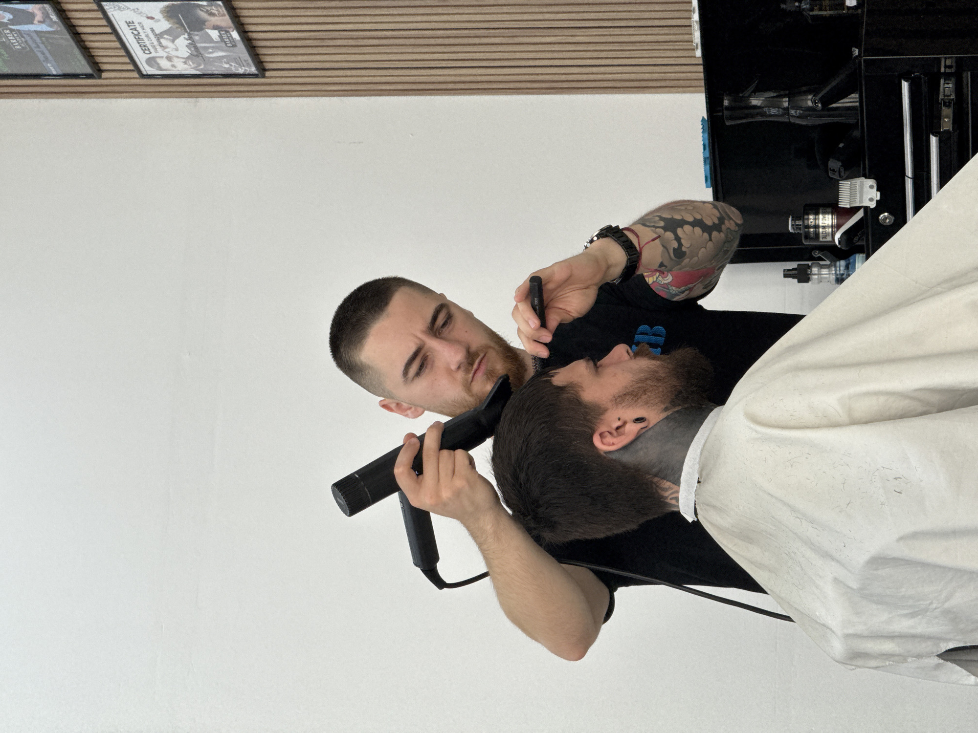 An indoor, eye-level shot captures a barber diligently working on a client's beard in a modern barbershop, likely located in Sofia, Bulgaria. The image is composed at an angle, showing the barber on the left and the client on the right, both focused downwards.
The barber, a man with short hair and a neatly trimmed beard, wears a dark short-sleeved shirt, revealing a heavily tattooed left arm with intricate, dark designs, and a watch on his wrist. He holds an electric hair trimmer in his left hand, carefully working on the client's jawline, and a smaller black tool, possibly for detailing, in his right hand. His expression is one of concentration.
The client, a man with dark hair and a substantial beard, is seated and covered by a white barber's cape, which is speckled with small hair trimmings. He has a dark tattoo on his neck and an ear piercing visible. He appears relaxed, with his head slightly tilted for the barber's access.
In the background, a clean white wall occupies most of the space. Above it, two framed certificates or pictures are visible, hanging beneath a wooden slatted panel. Partial text on one frame reads "CERTIFICATE" and "BARBER". To the right, behind the client, a dark barber station or cabinet holds various tools and products, including a bottle labeled "TITANIUM POWER". The lighting is bright and even, indicating an indoor setting during daytime hours.
