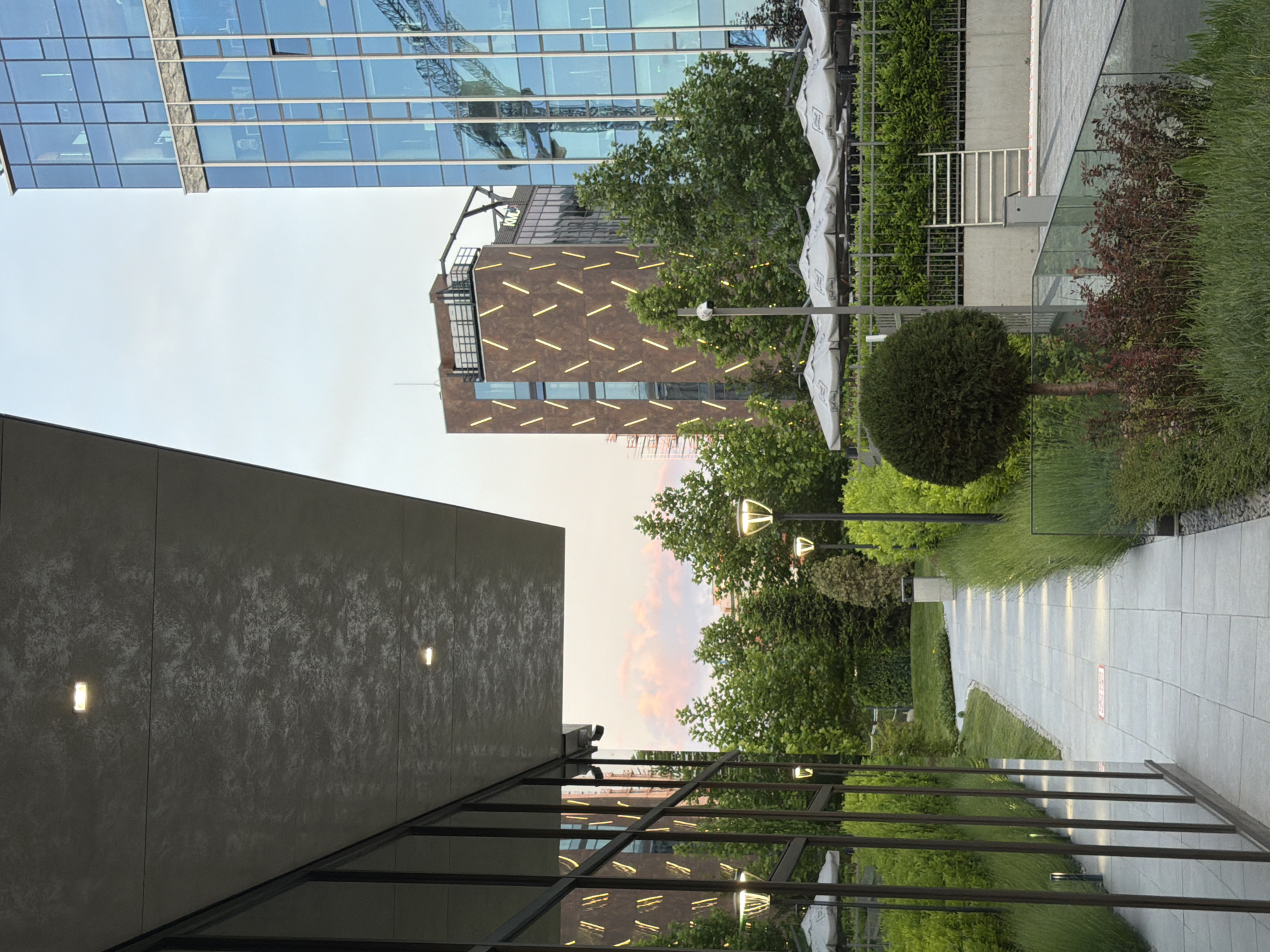 The image depicts a modern urban outdoor area in Sofia, Bulgaria, likely a courtyard or plaza between contemporary buildings. The foreground features a pathway paved with light-colored square tiles, bordered by lush green grass, manicured hedges, and ornamental trees. Tall, slender modern lampposts illuminate the walkway. To the left, a glass-fronted building reflects the surrounding environment, with a large, angled concrete overhang providing shade. In the midground, a curved path leads towards a cluster of trees. Behind the greenery, a row of white umbrellas shades an outdoor seating area, protected by a metal fence. Further back, a distinctive cylindrical building with a dark facade is adorned with diagonal, illuminated golden lines, and a construction crane is visible. Dominating the right side of the frame is a towering glass skyscraper, its facade reflecting the sky and the adjacent crane. The sky is clear with soft, pastel-colored clouds, suggesting either dawn or dusk. No people are present in the scene.