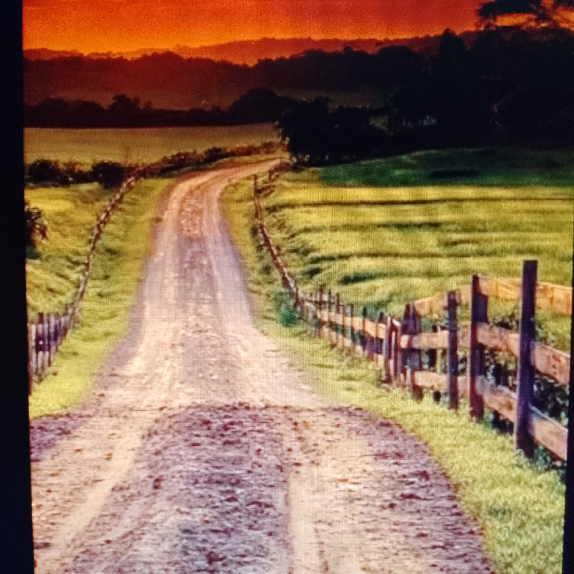 A dirt road winds through a rural landscape under a vivid orange sunset. The road is flanked by green fields and wooden fences. In the background, there are silhouetted hills and trees.