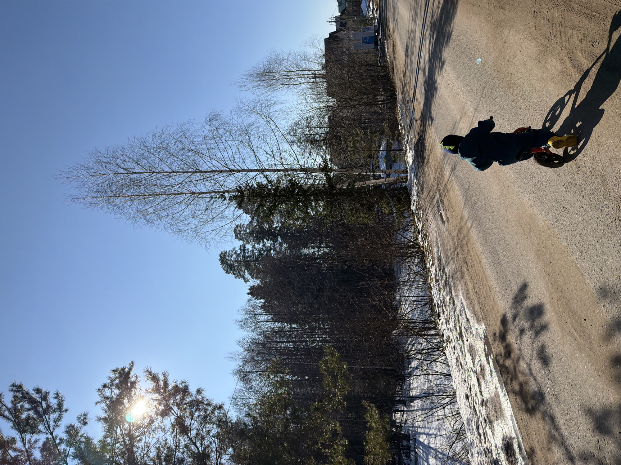 A young child on a bicycle is riding on a paved road. The child is wearing dark clothing and yellow shoes. The road is lined with trees and snow on one side. The sky is blue and sunny. The location is Chaikovsky, Russia. The presence of snow suggests it is winter or early spring.