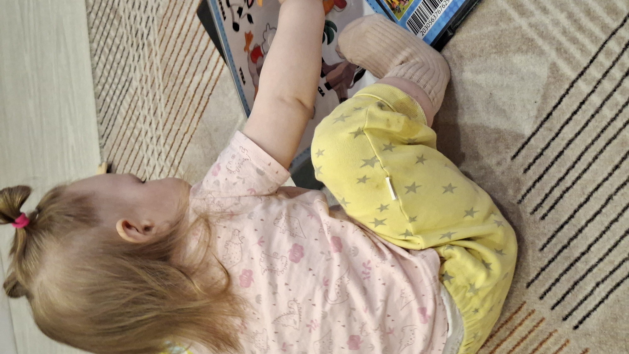 A young child lies on a patterned rug, wearing a pink dinosaur-patterned top and yellow star-patterned bottoms with beige socks. They are holding a book open with one hand. The child's hair is blonde and pulled into a small ponytail with a pink hair tie. A portion of a barcode is visible on the book. The location and city are unknown.